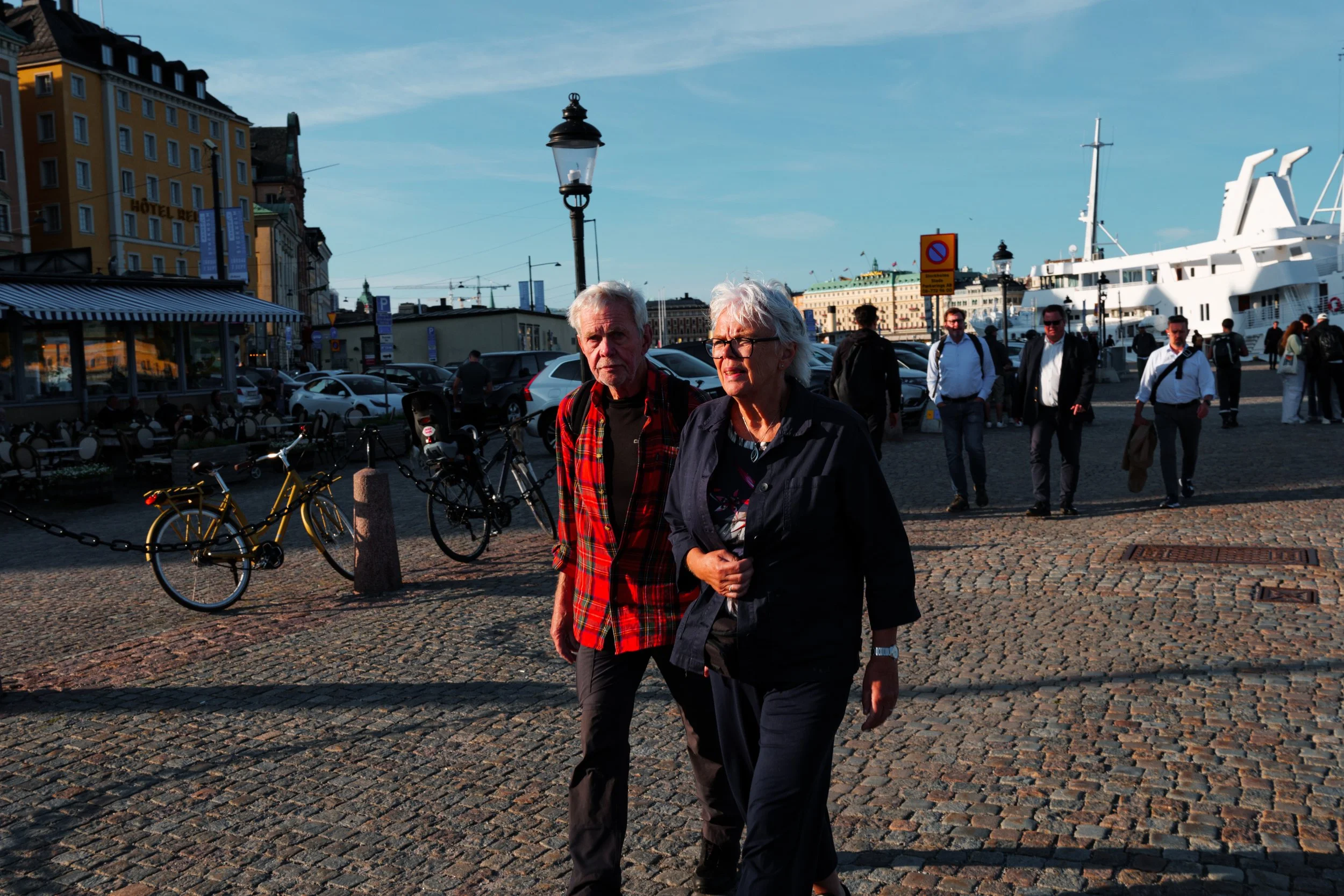Older man and woman walking on cobblestone street near harbor with boats and people in background, under clear blue skies.