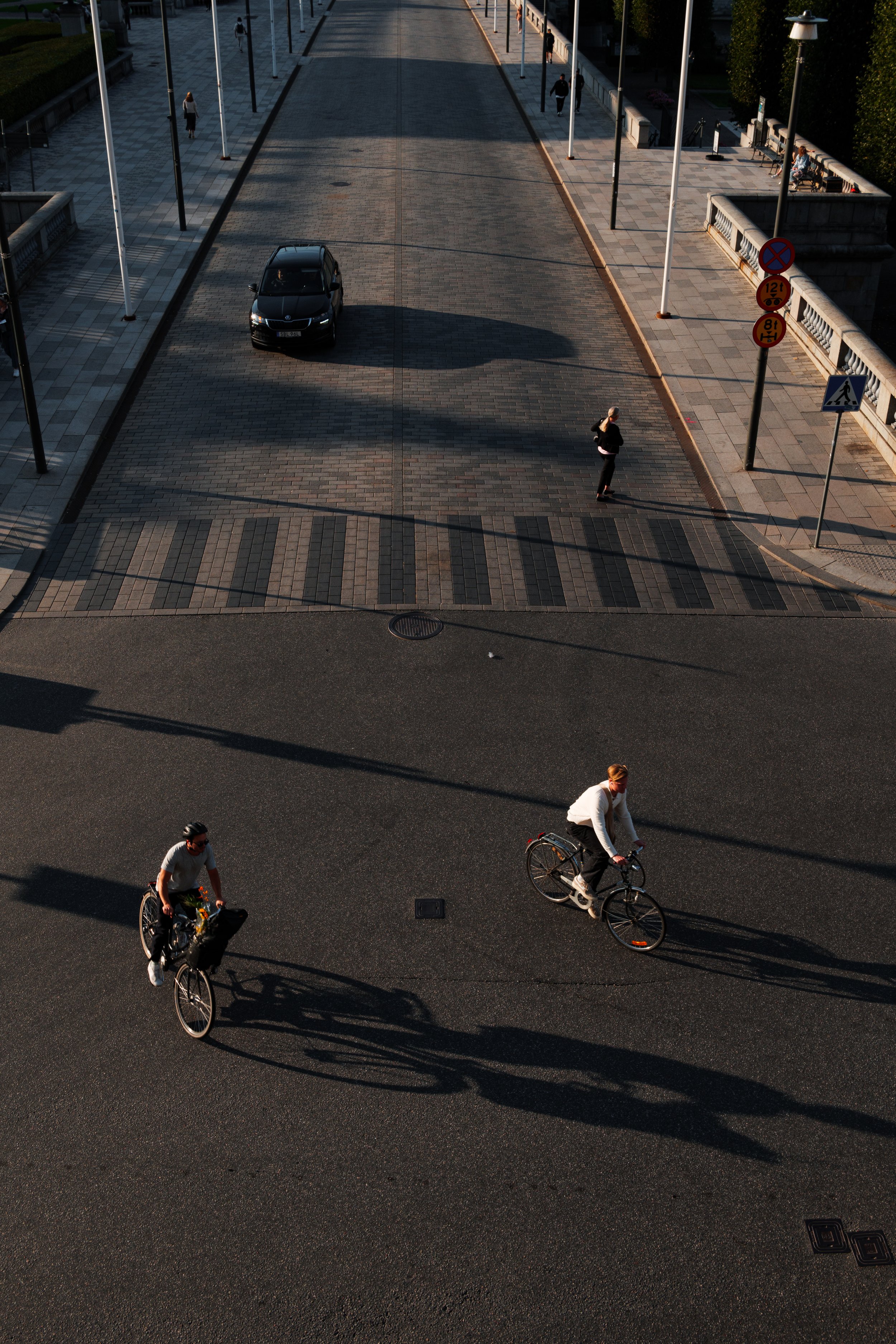 Two people riding bicycles on a dark road, casting long shadows. One person with a black helmet and a backpack, the other with a white shirt. In the background, a woman is walking across a crosswalk near a parked car and street signs. The street is l