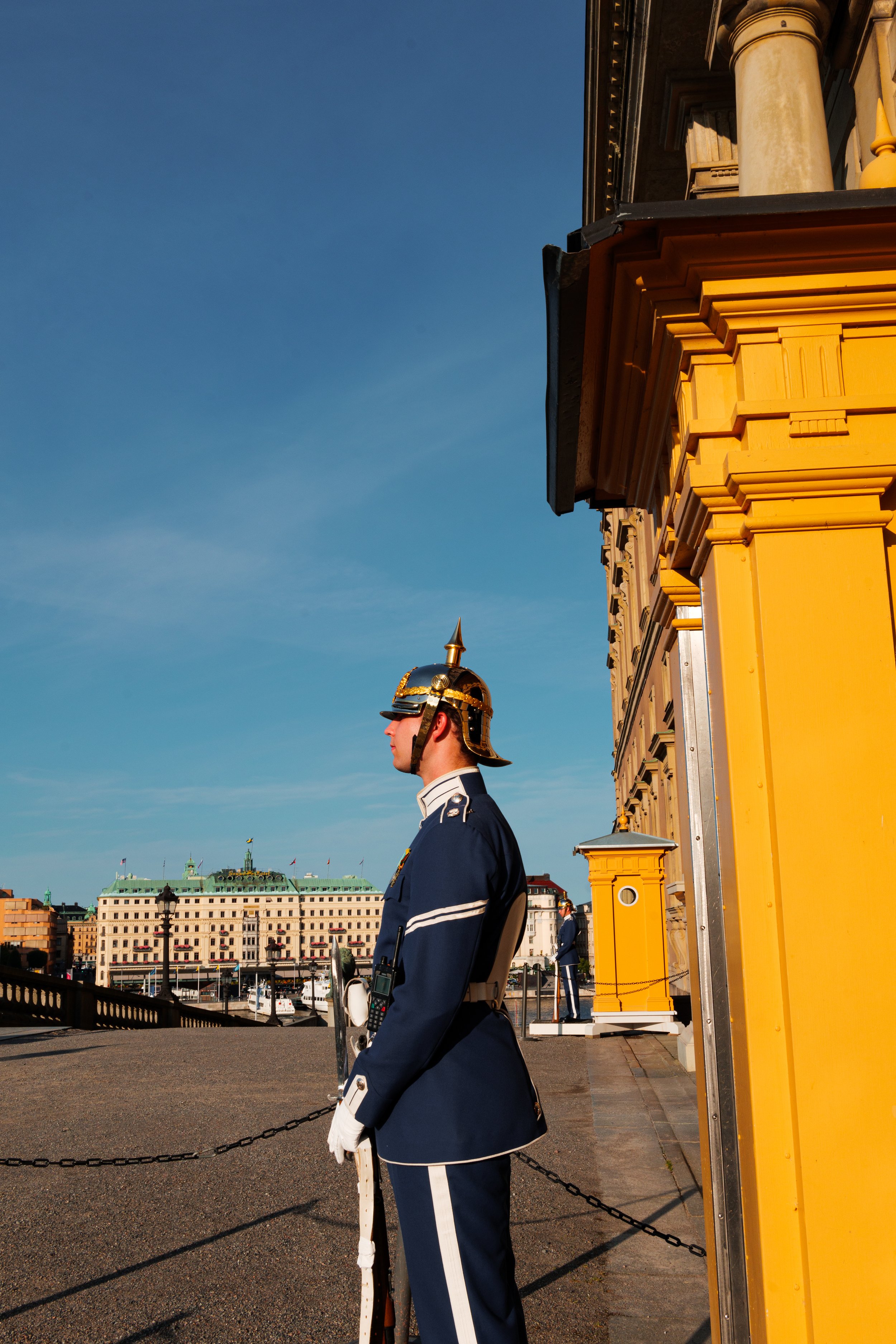 A uniformed guard standing with a helmet, holding a rifle, in front of a yellow building, with a cityscape and a large white building with green roof in the background.