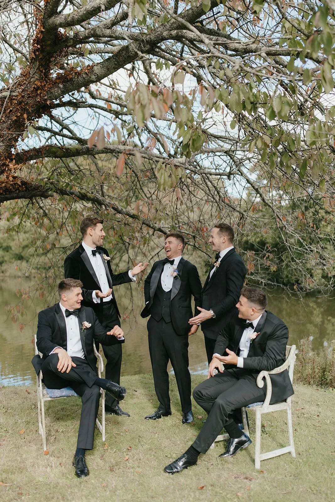 Six men in tuxedos with bow ties, some seated on chairs and others standing, laughing and talking outdoors near a lake with a large tree overhead.