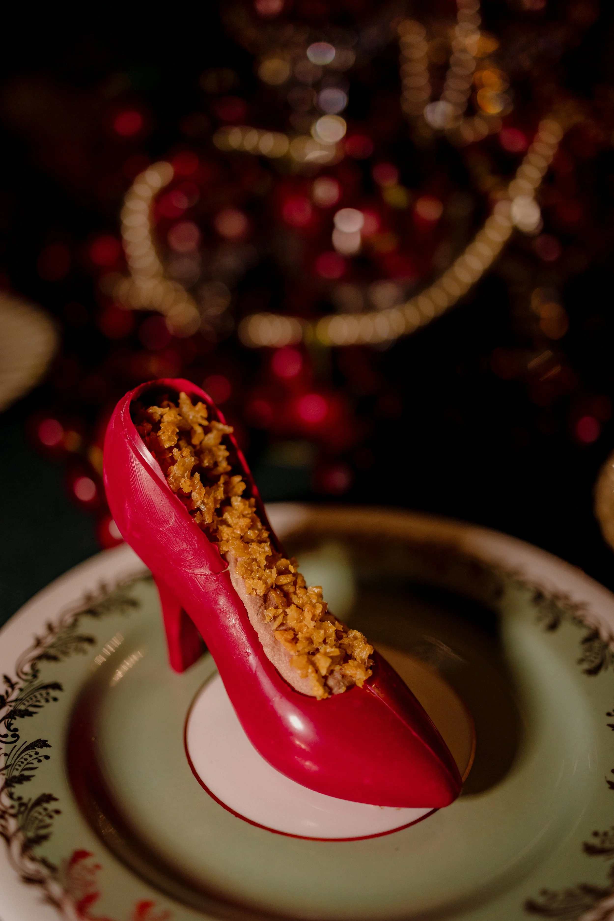 A red high heel shoe filled with a crumbly pastry, placed on a decorative plate, with blurred holiday lights in the background.