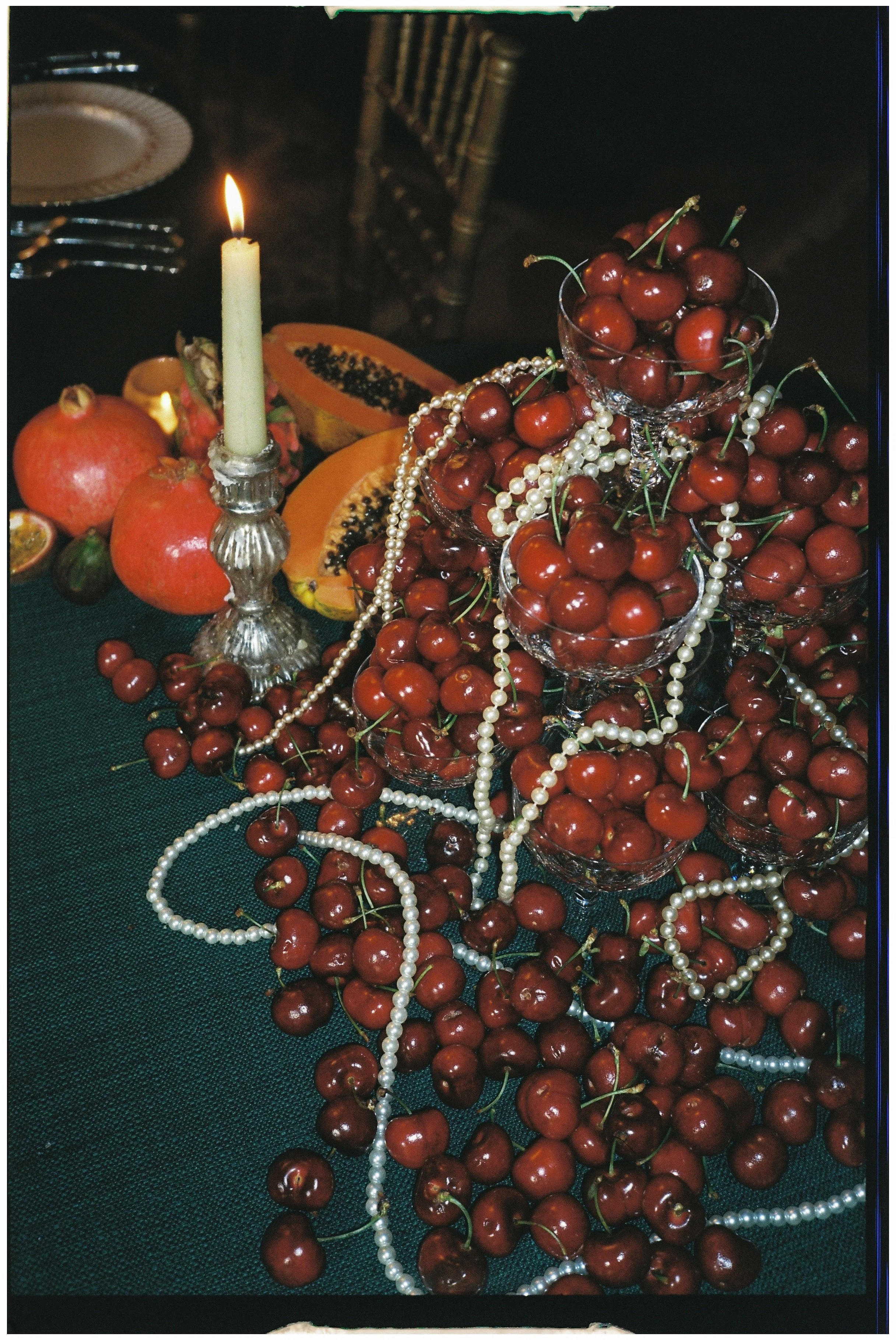 A table decorated with pomegranates, halved papayas, and pearls, with a lit candle in a silver holder in the background.