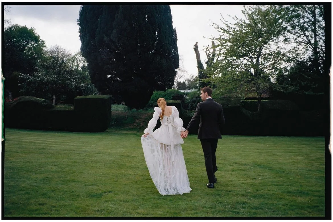 A couple walking hand in hand on a grassy lawn, with the woman in a white wedding dress and the man in a dark suit, in a lush outdoor setting.