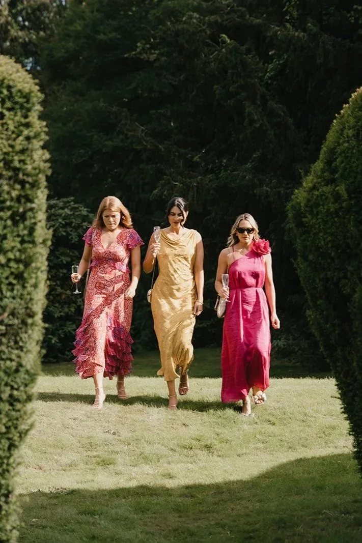 Three women in elegant dresses walking on a grassy lawn holding champagne glasses, surrounded by tall green bushes and trees.