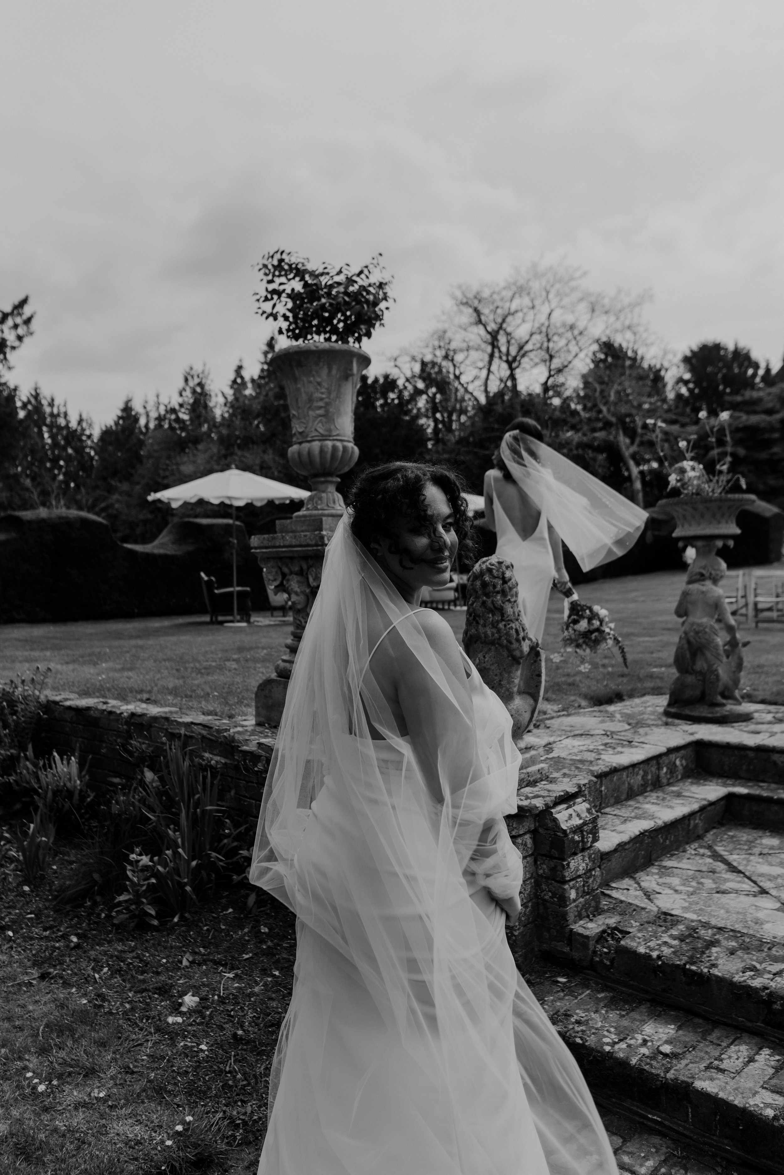 A woman in a wedding dress and veil smiling at the camera outdoors near steps with sculptures and garden features in the background.