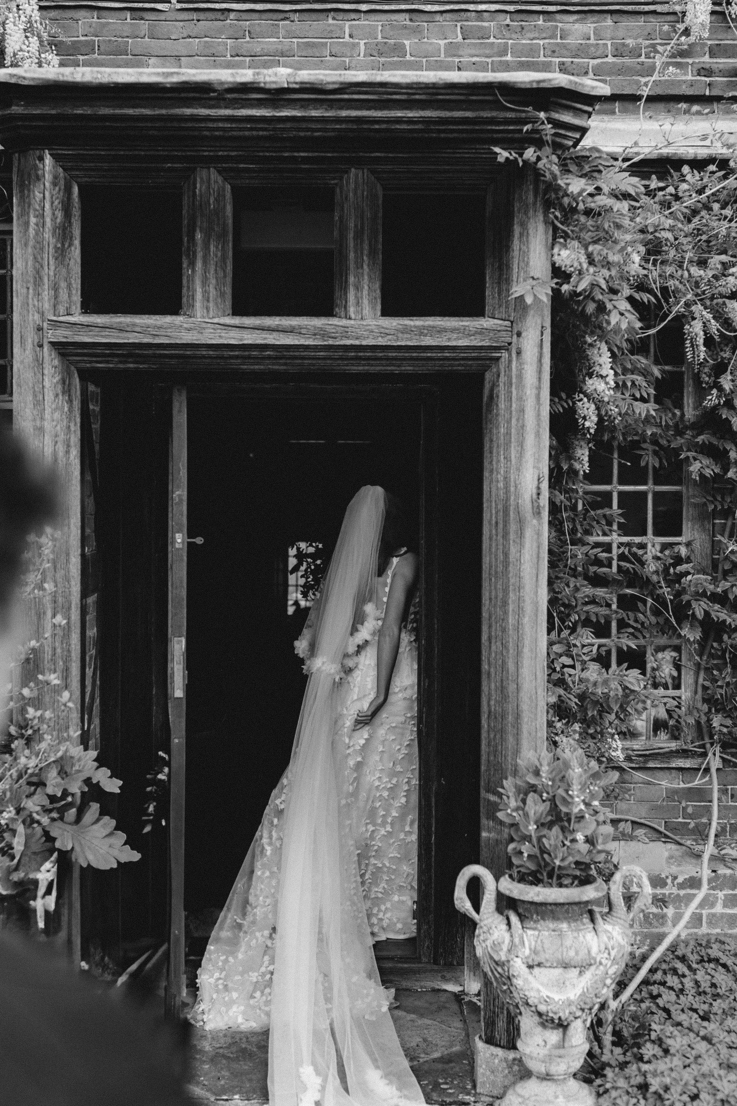 A black and white photo of a bride in a wedding dress standing in a doorway with a long veil covering her face. The scene includes an outdoor garden setting with a potted plant and leafy vines.