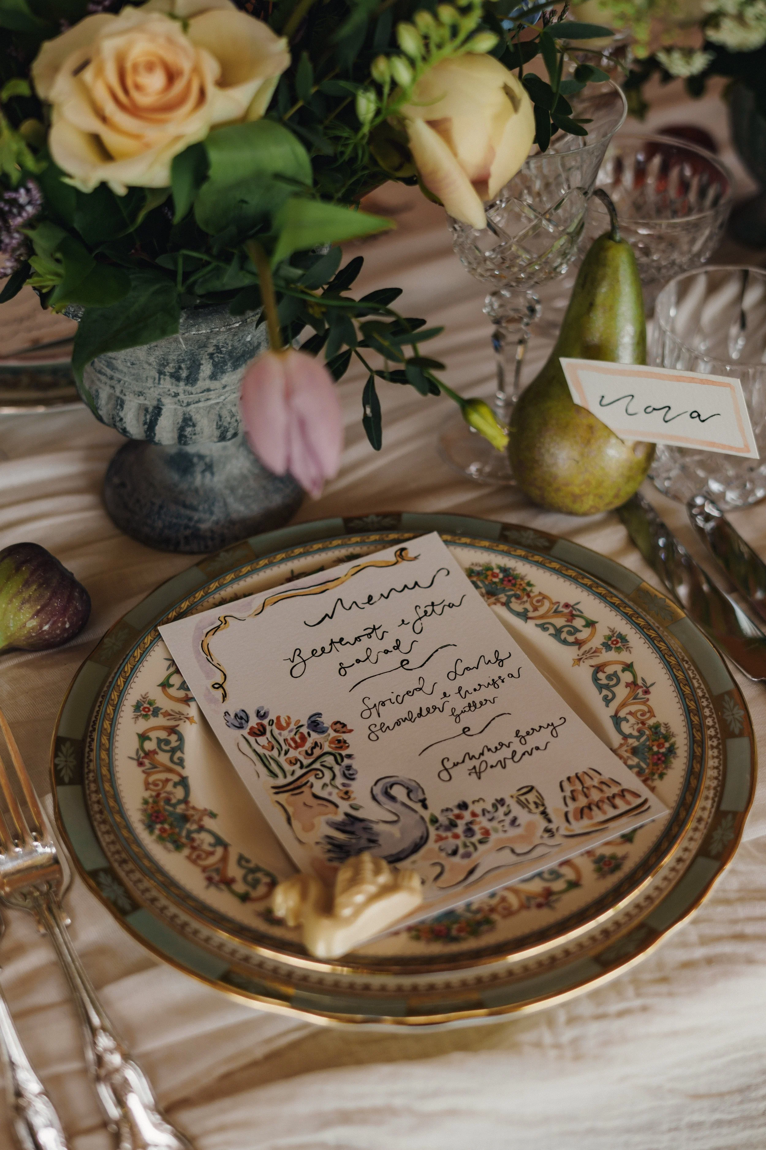 A decorated table setting with a floral centerpiece, ceramic plate with a handwritten menu, a pear, a name card, and cutlery. The menu features colorful illustrations including a swan, flowers, and a basket, indicating a formal or rustic event.