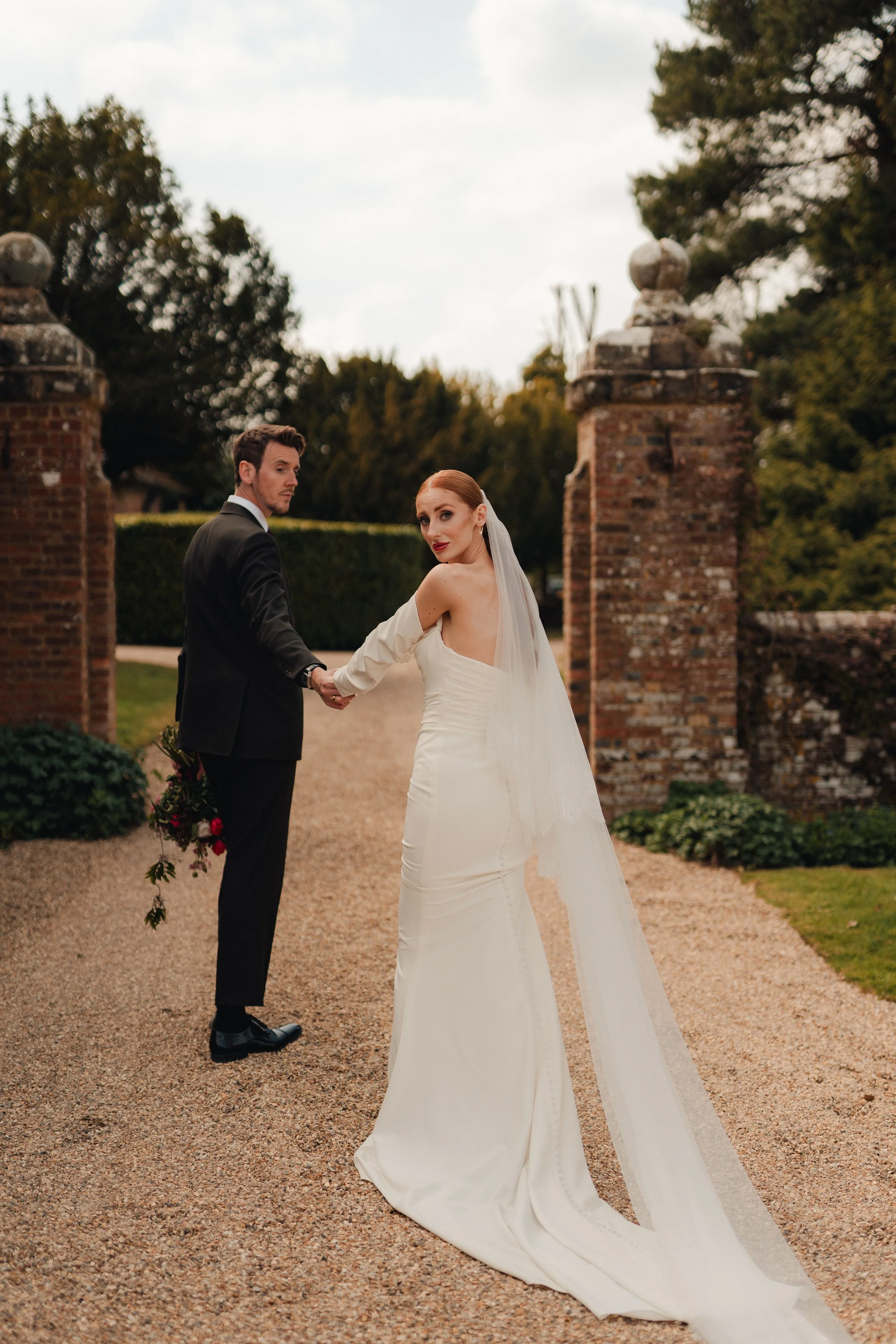 A bride and groom holding hands outdoors, with dark green trees and stone pillars in the background, on a gravel pathway.