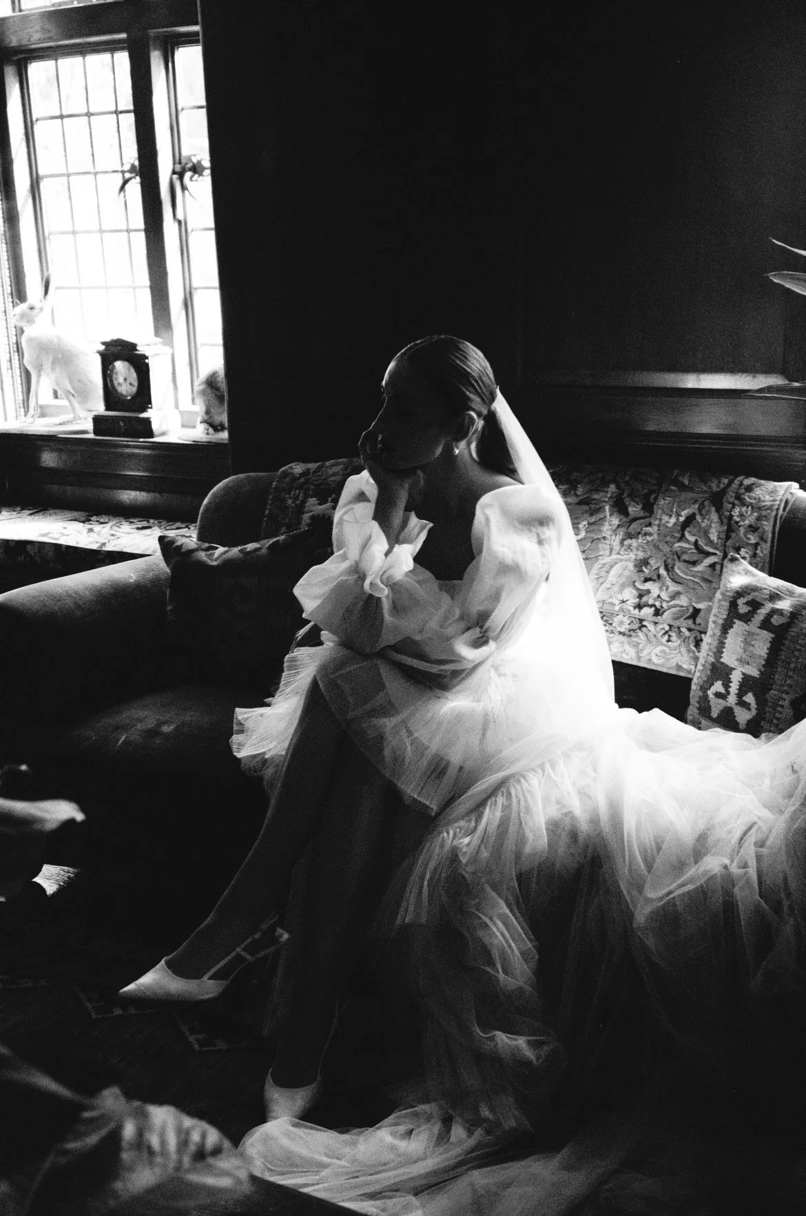 A woman in a wedding dress sitting on a sofa in a dimly lit room with a window, two cats on the windowsill, and decorative pillows.