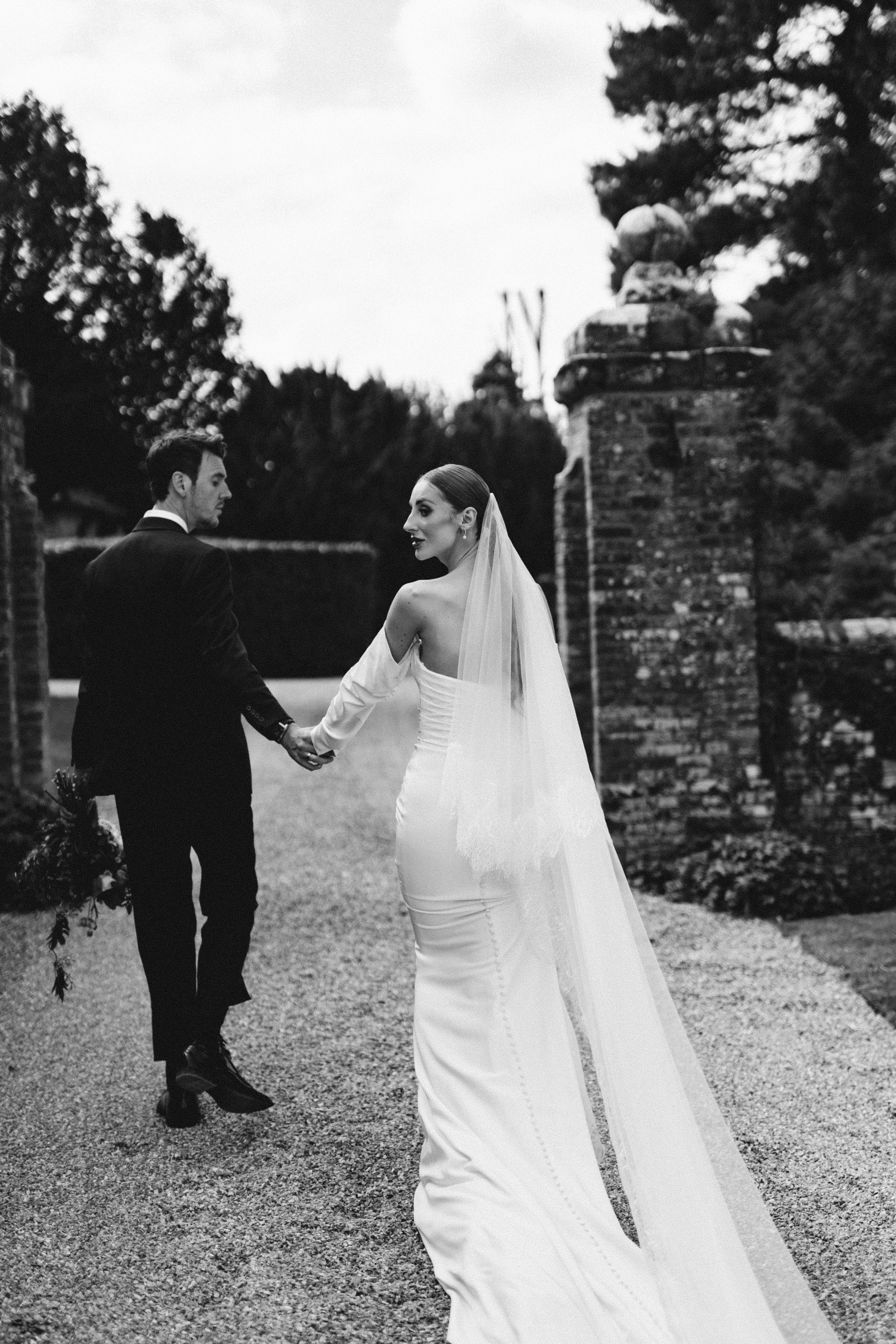 Black and white photo of a bride and groom holding hands outdoors. The bride wears a long, strapless wedding dress and veil, and the groom wears a suit. They are walking along a gravel path between brick pillars and trees.