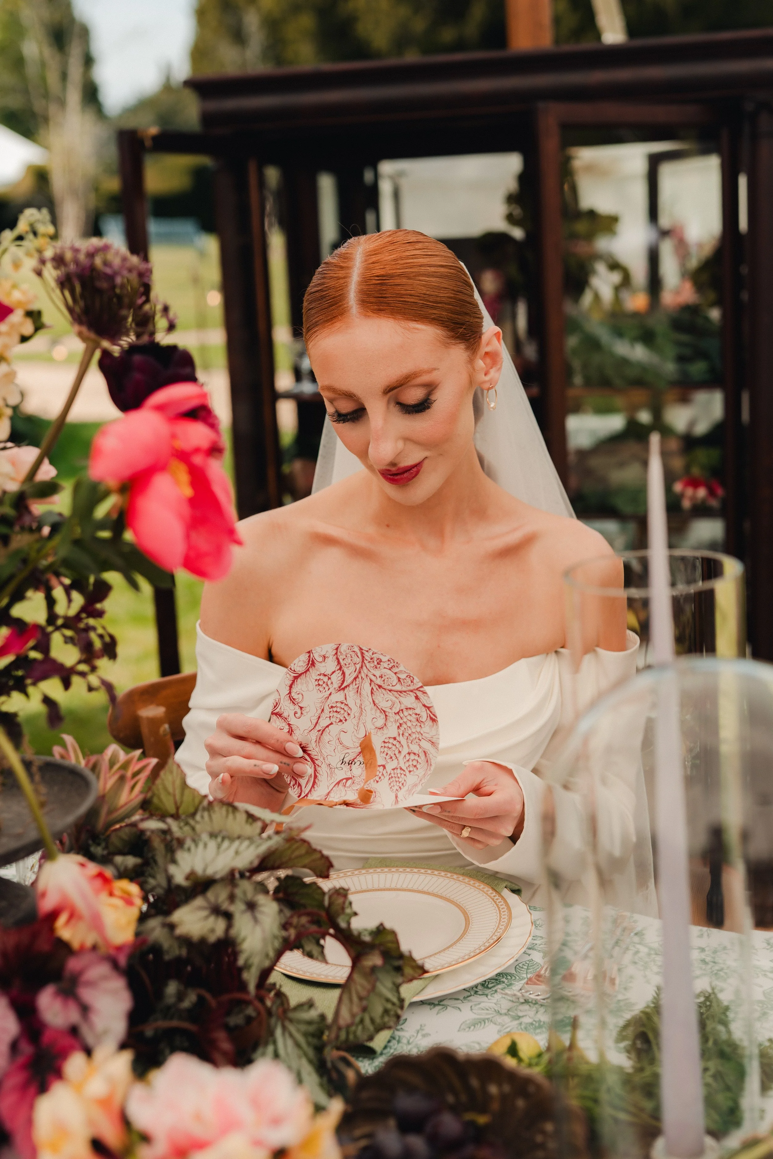 A bride with red hair and makeup, wearing an off-shoulder white wedding dress and pearl earrings, sits at a table decorated with flowers and greenery, holding a paper napkin. The setting appears to be an outdoor wedding reception with a wooden garden shed and plants in the background.