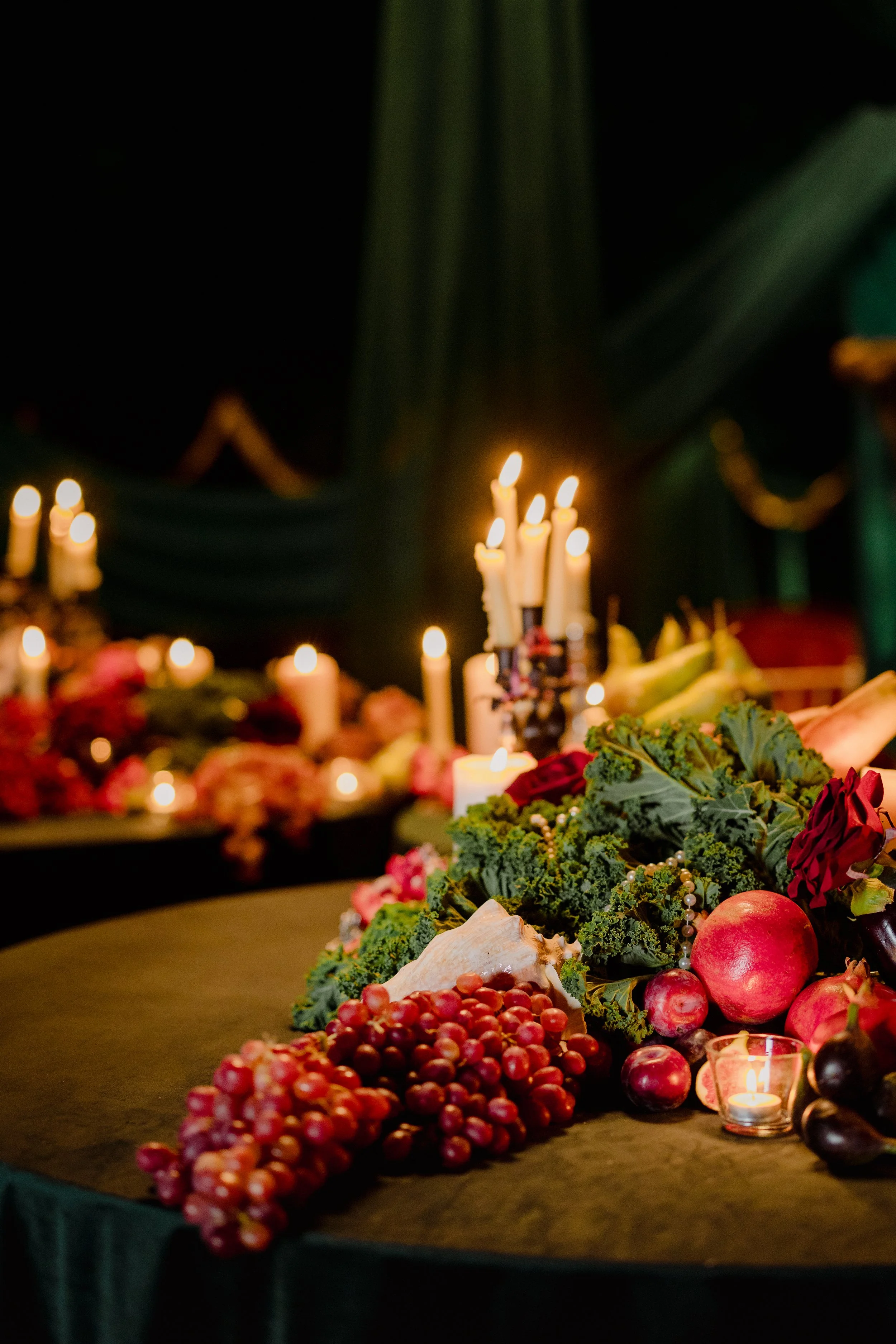 A table with various fruits like grapes, pomegranates, and eggplants, decorated with greenery, candles, and flowers, set against a dark background with lit candles
