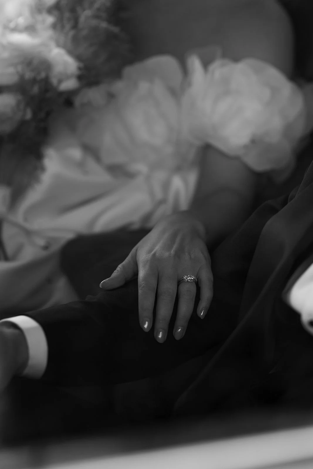 A bride and groom holding hands, with the bride's hand resting on the groom's arm, showing her wedding ring, in a black and white photo.