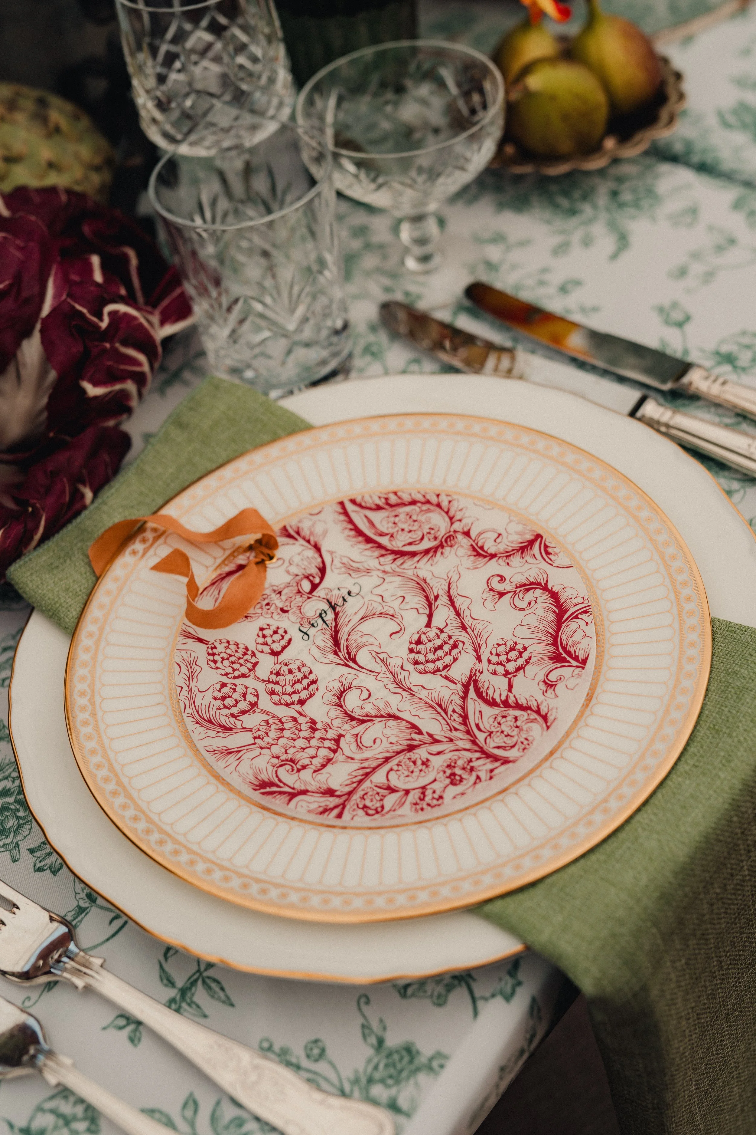 A set dining table with ornate plates, green napkin, cutlery, crystal glasses, a bowl of pears, and decorative elements.