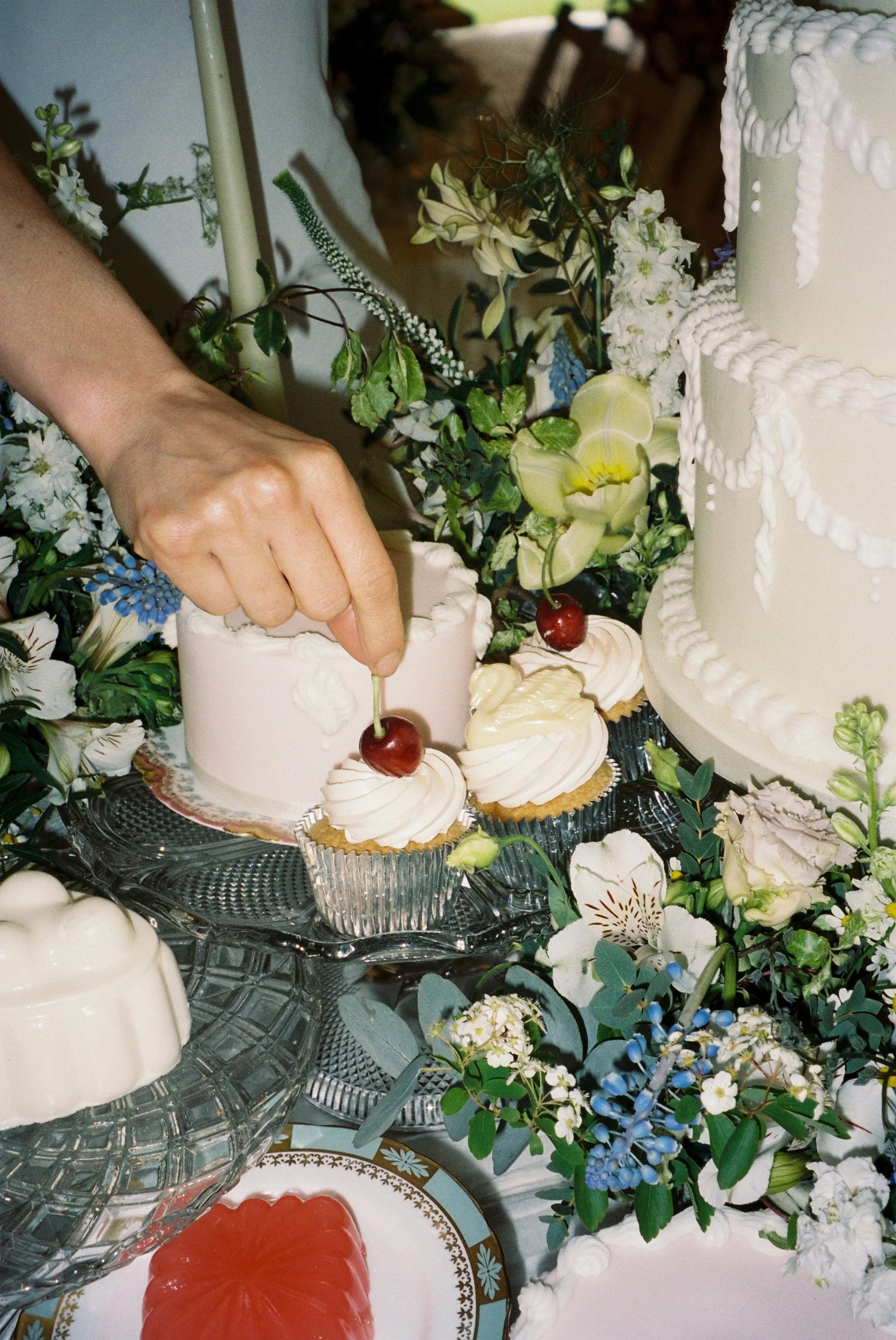 A person's hand decorates a wedding cake with two cherries on cupcakes, surrounded by flowers and other desserts.