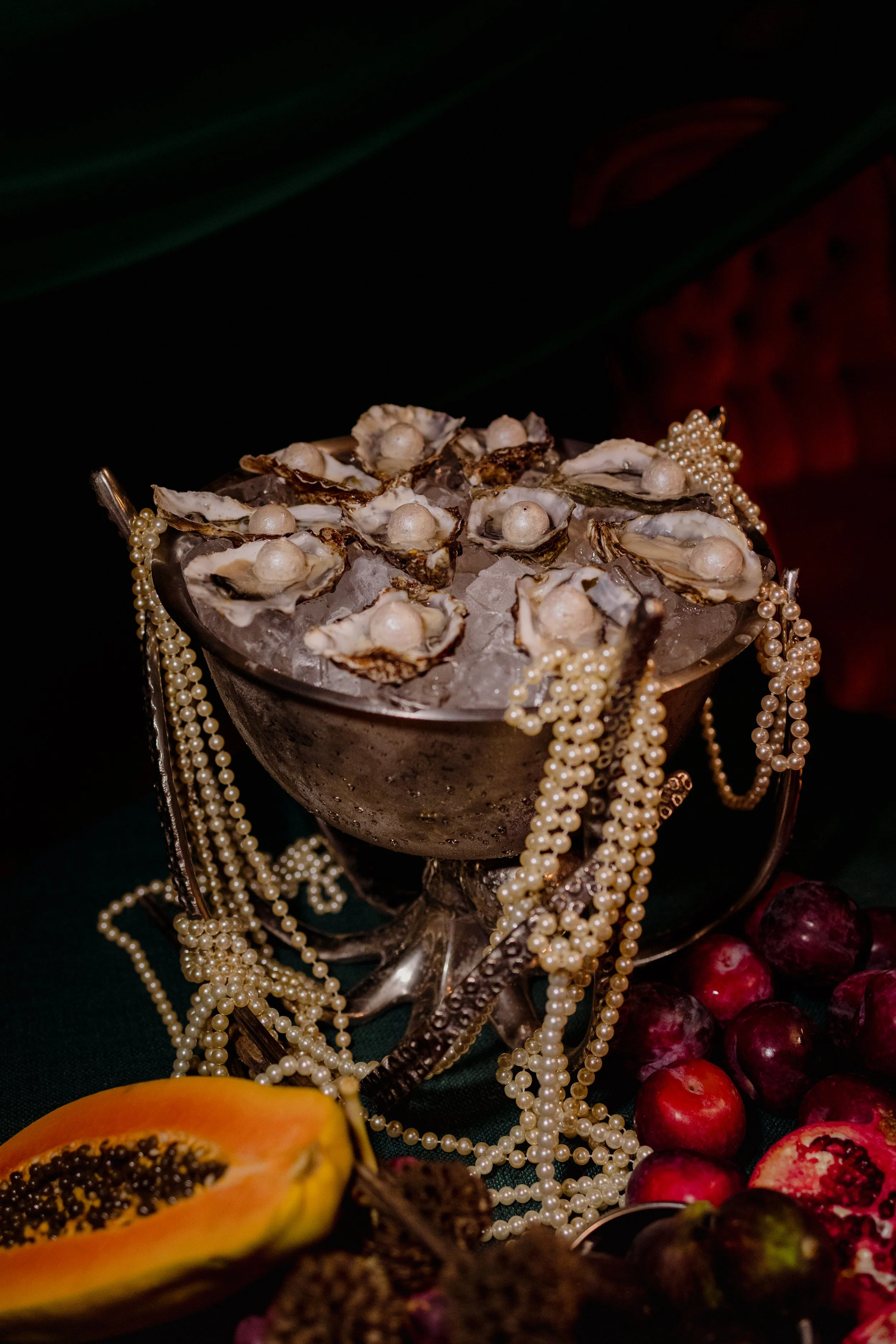 A silver bowl filled with oysters on ice, adorned with pearl necklaces, with fruits including a halved papaya and grapes surrounding it.