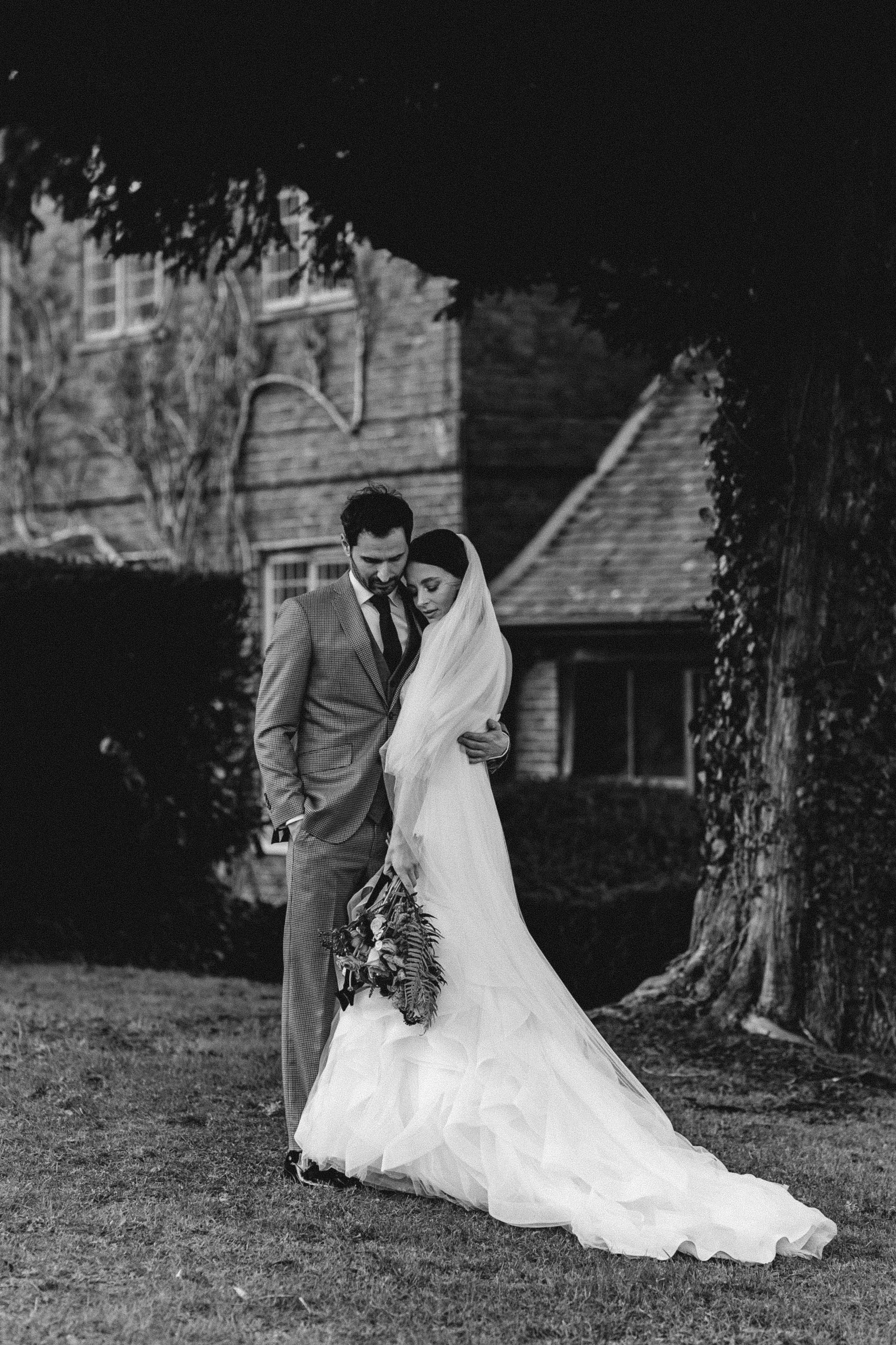 Black and white photo of a bride and groom standing close together outdoors, with the bride holding a bouquet of flowers and both looking down, in front of a large tree and a house in the background.