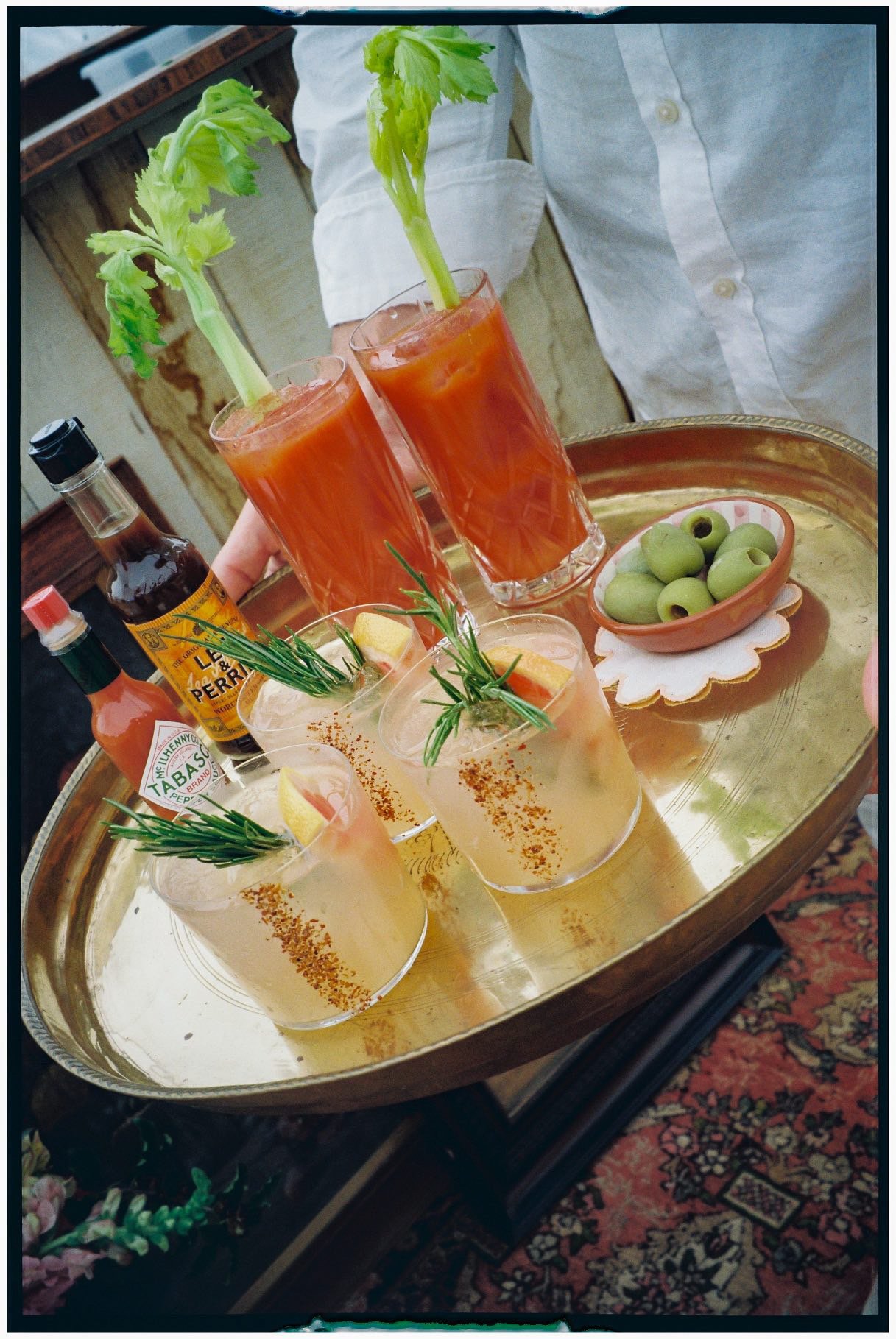A tray of drinks with celery stalks, olives, hot sauce, and whiskey, garnished with lemon and sprigs of rosemary, alongside a bowl of green olives.