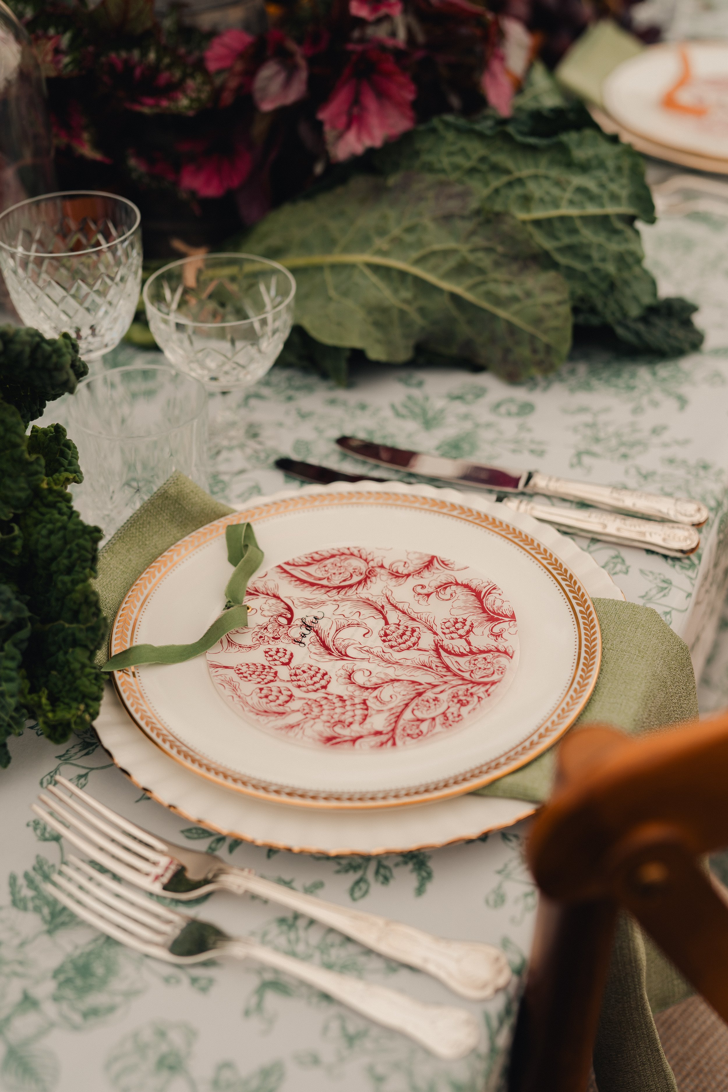 A beautifully set dining table with a patterned tablecloth, a decorative plate with a red floral design, silverware, empty crystal glasses, and green leaves as part of the table decor.