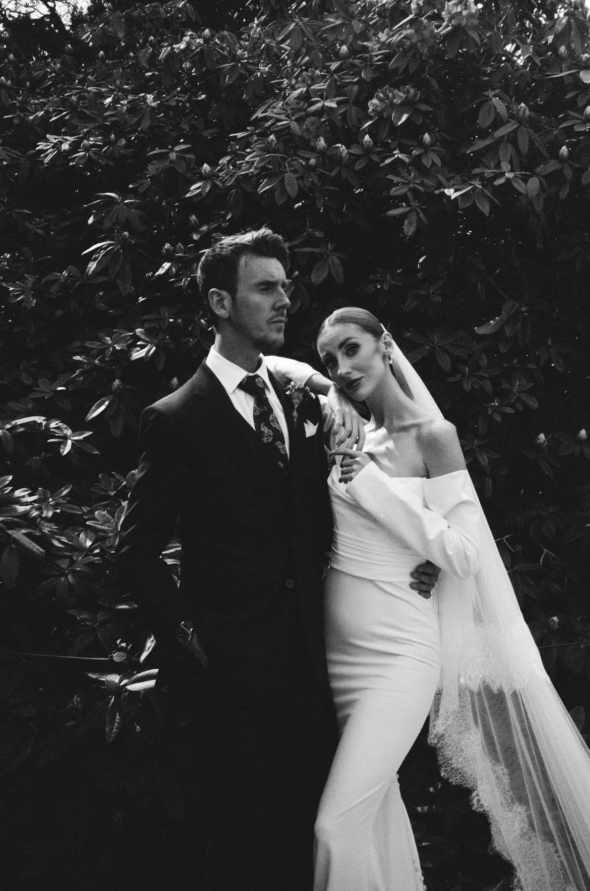 Black and white photo of a couple in wedding attire standing close to each other outdoors with dense foliage in the background. The man is wearing a suit, and the woman is dressed in a white off-shoulder gown with a veil.
