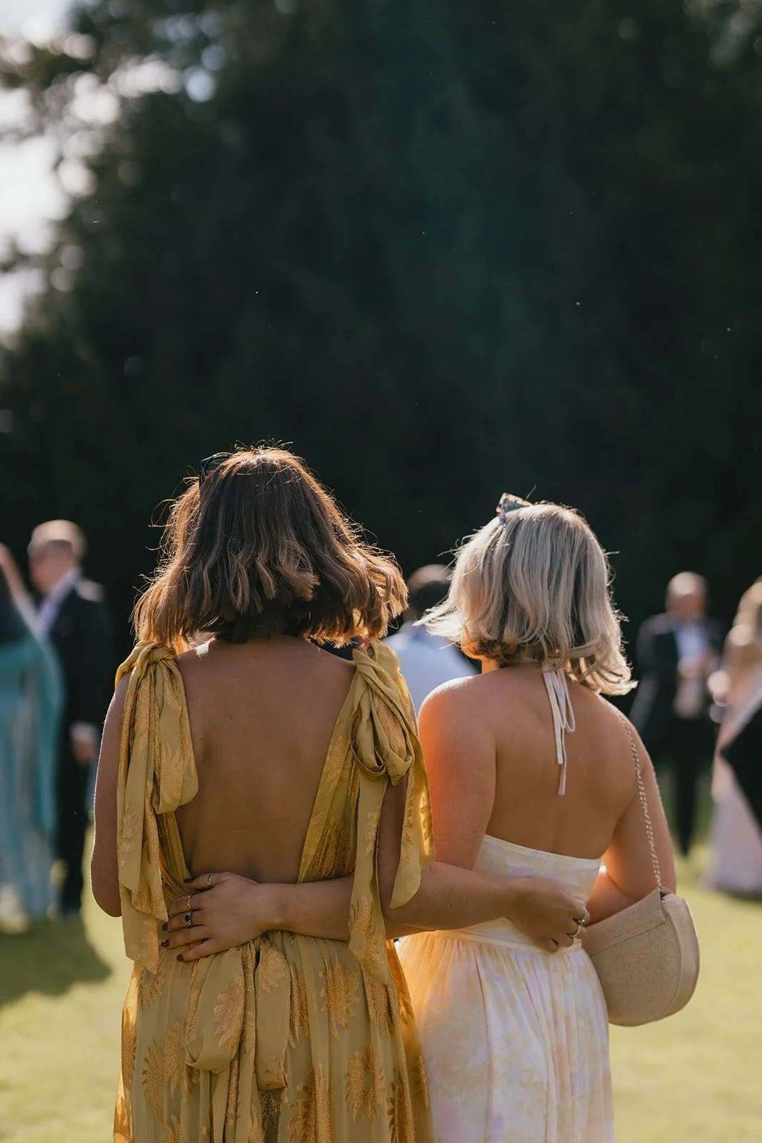 Two women with shoulder-length hair, one with dark brown hair and the other with blonde hair, wearing sleeveless dresses, standing outdoors at a social event, back view.