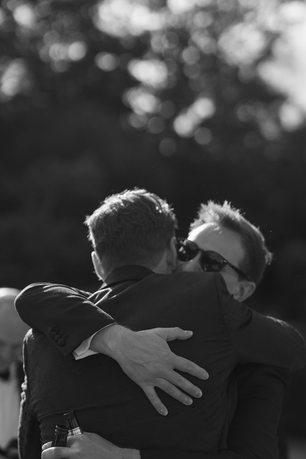 Two men in suits embracing outdoors, one wearing sunglasses, a beer bottle in hand, with sunlit trees in the background.