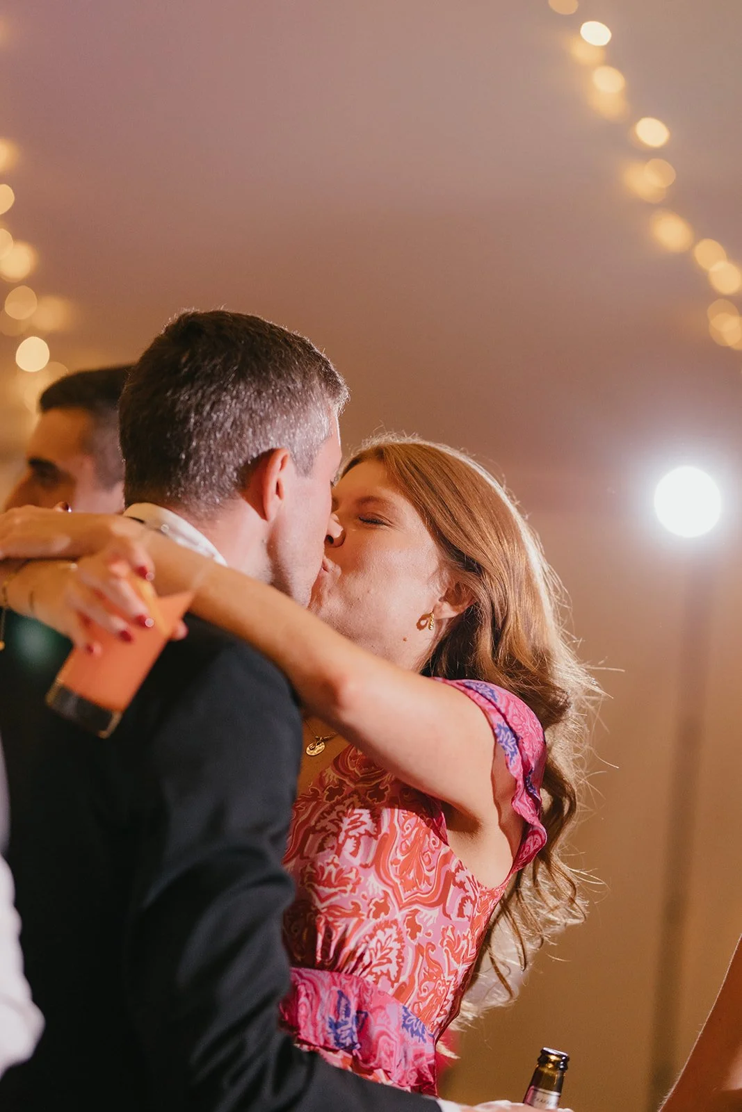 A woman with red hair and a woman with dark hair kiss at a celebration, with string lights visible in the background.