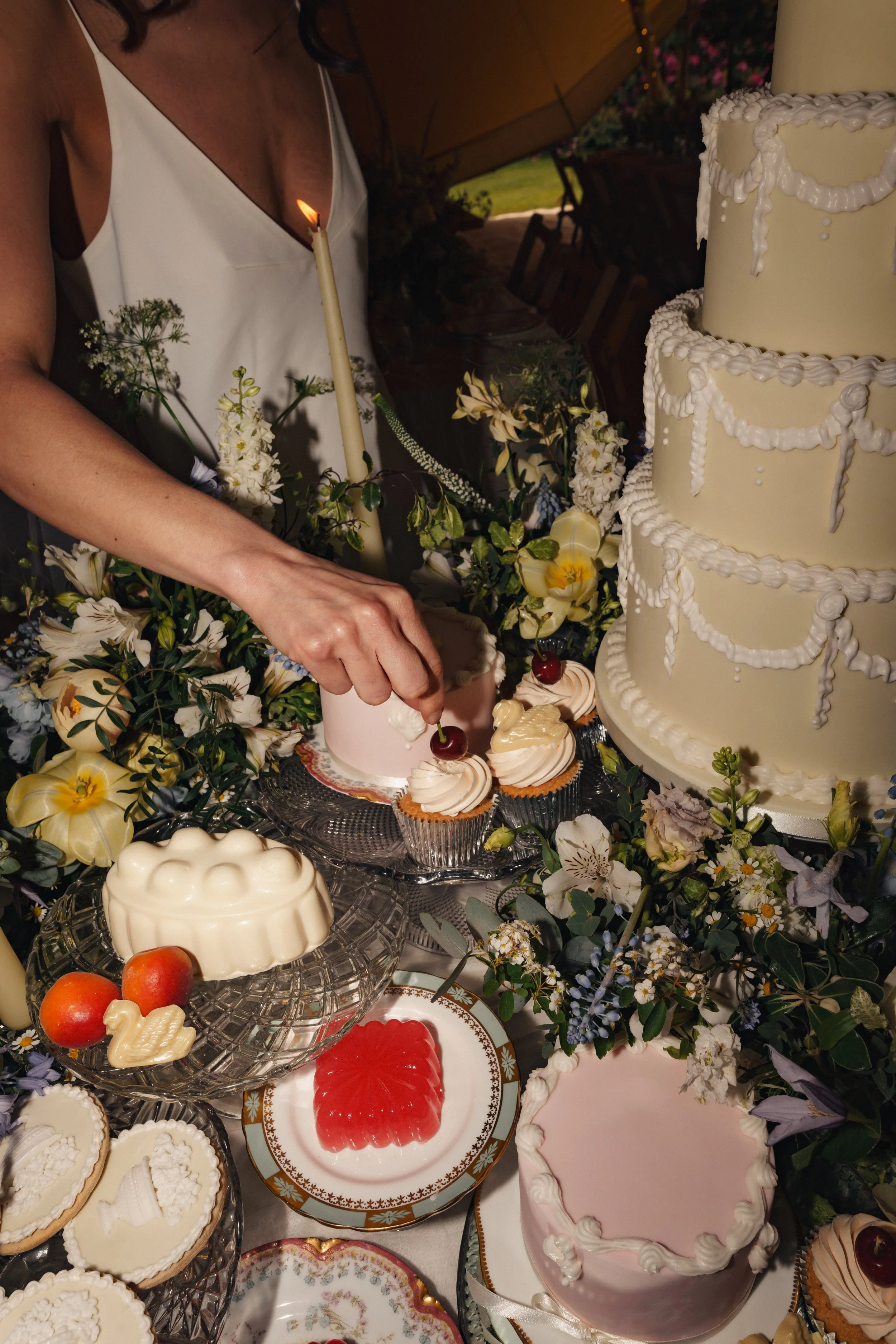 A woman in white dress decorating cupcakes at a wedding cake table, which features a large layered white cake with white icing decorations, surrounded by flowers, fruits, and various desserts.