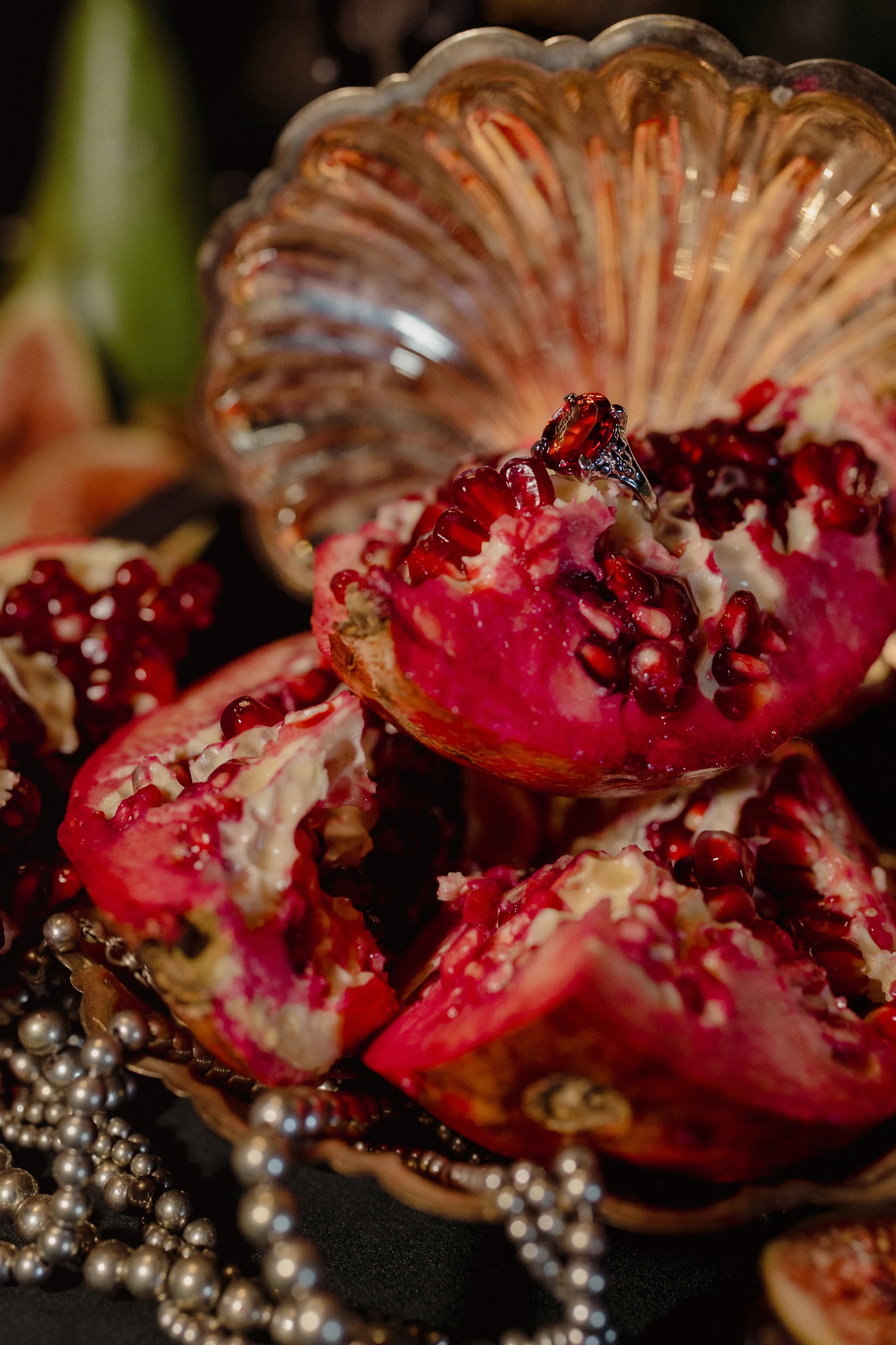 Close-up of a pomegranate with its seeds exposed, accompanied by a silver jewelry ring with a red gemstone, and strands of silver beads, in a decorative setting.