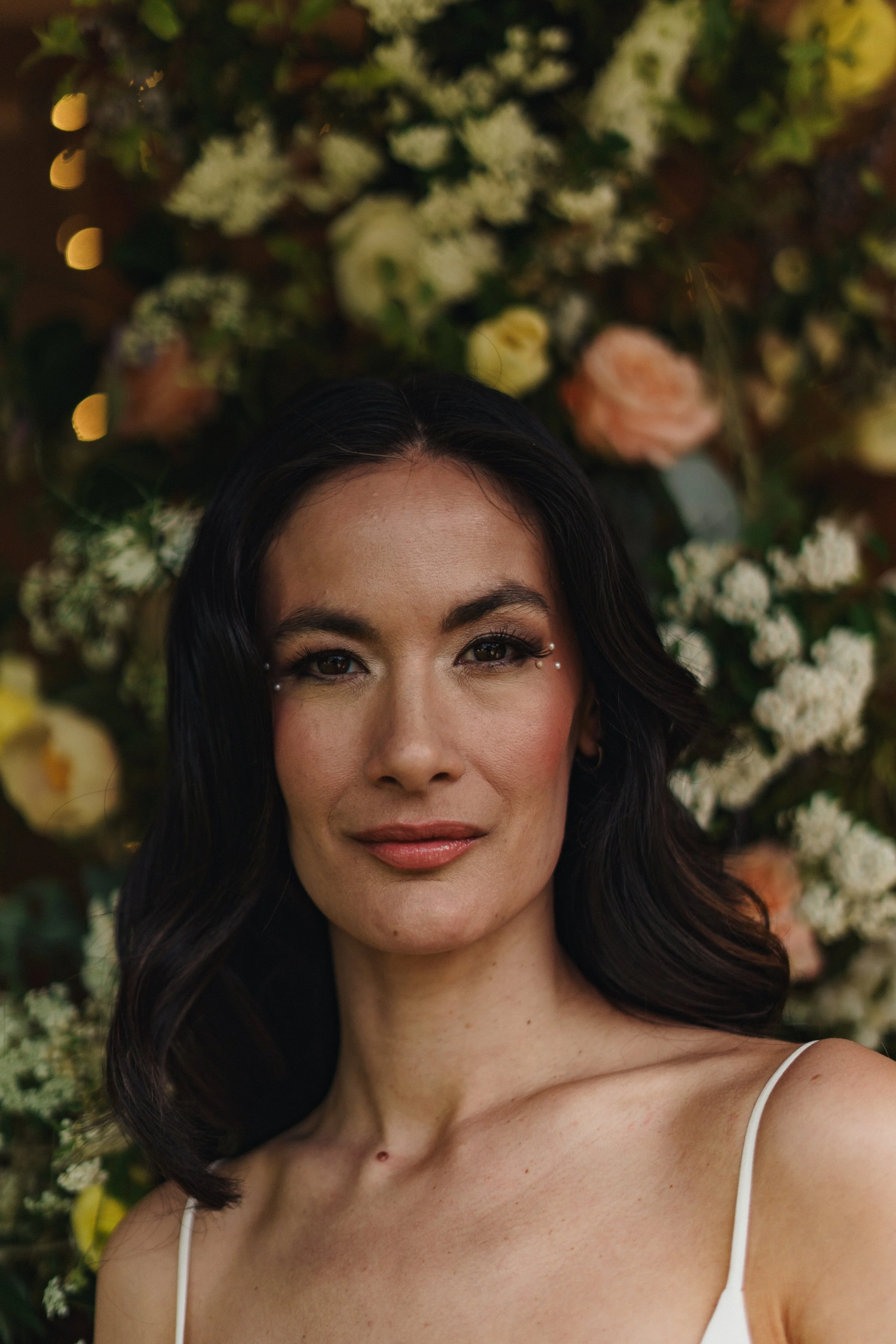 A woman with dark hair and makeup, wearing a white spaghetti strap top, stands in front of a floral background with white and pink roses, and small white flowers.