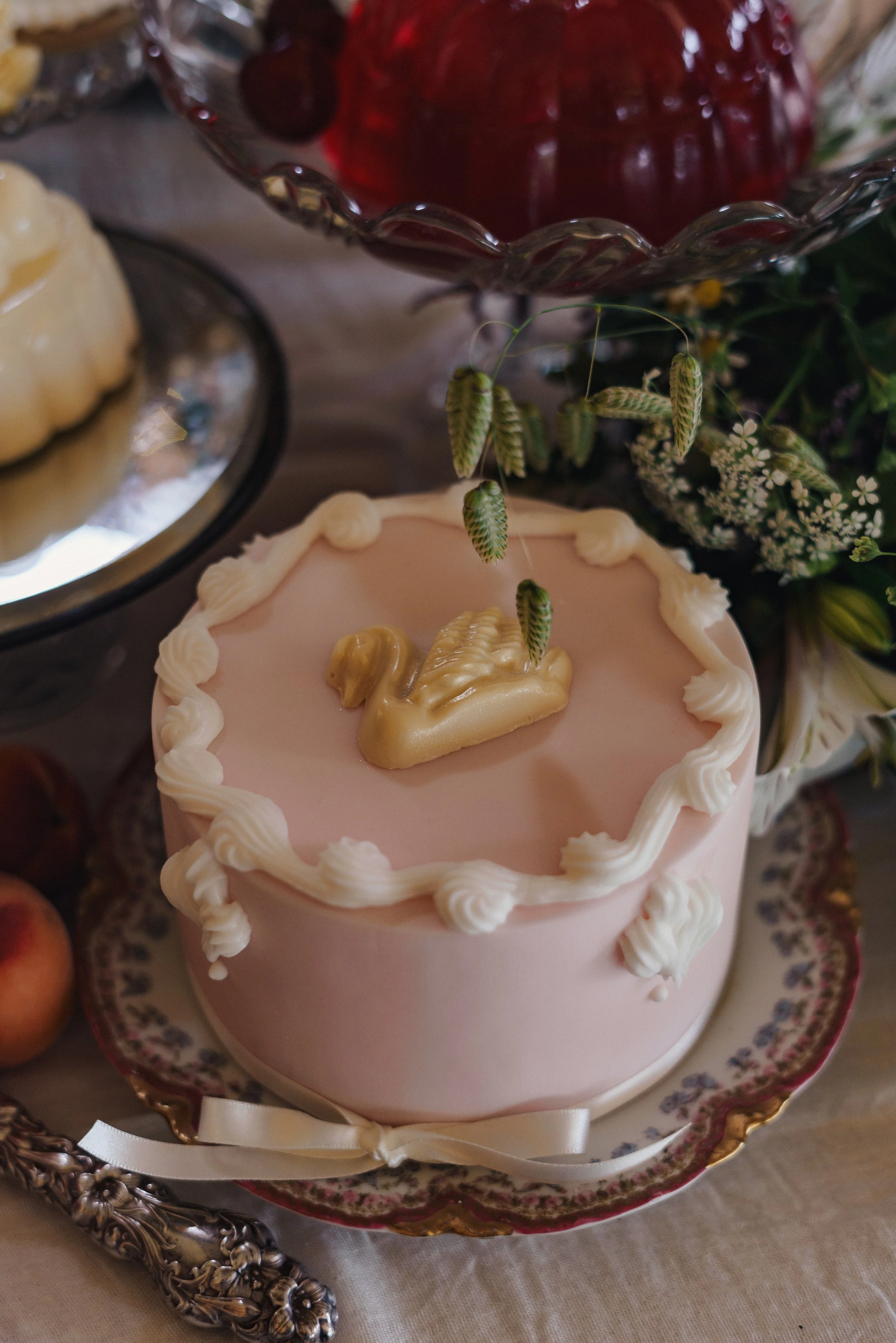 A pink cake with a white border on top, decorated with a gold swan and green wheat stalks, placed on a decorative plate, surrounded by flowers and other desserts.