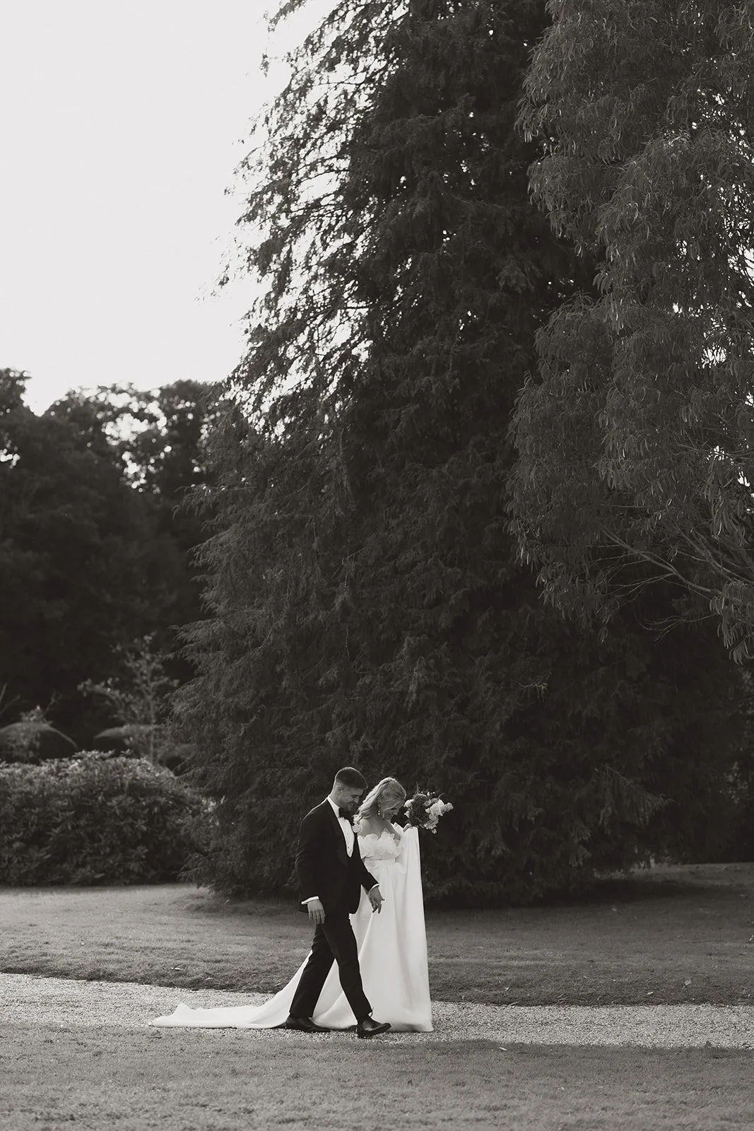A black and white photo of a bride in a long wedding dress holding a bouquet, and a groom in a suit walking together outdoors near a large tree.