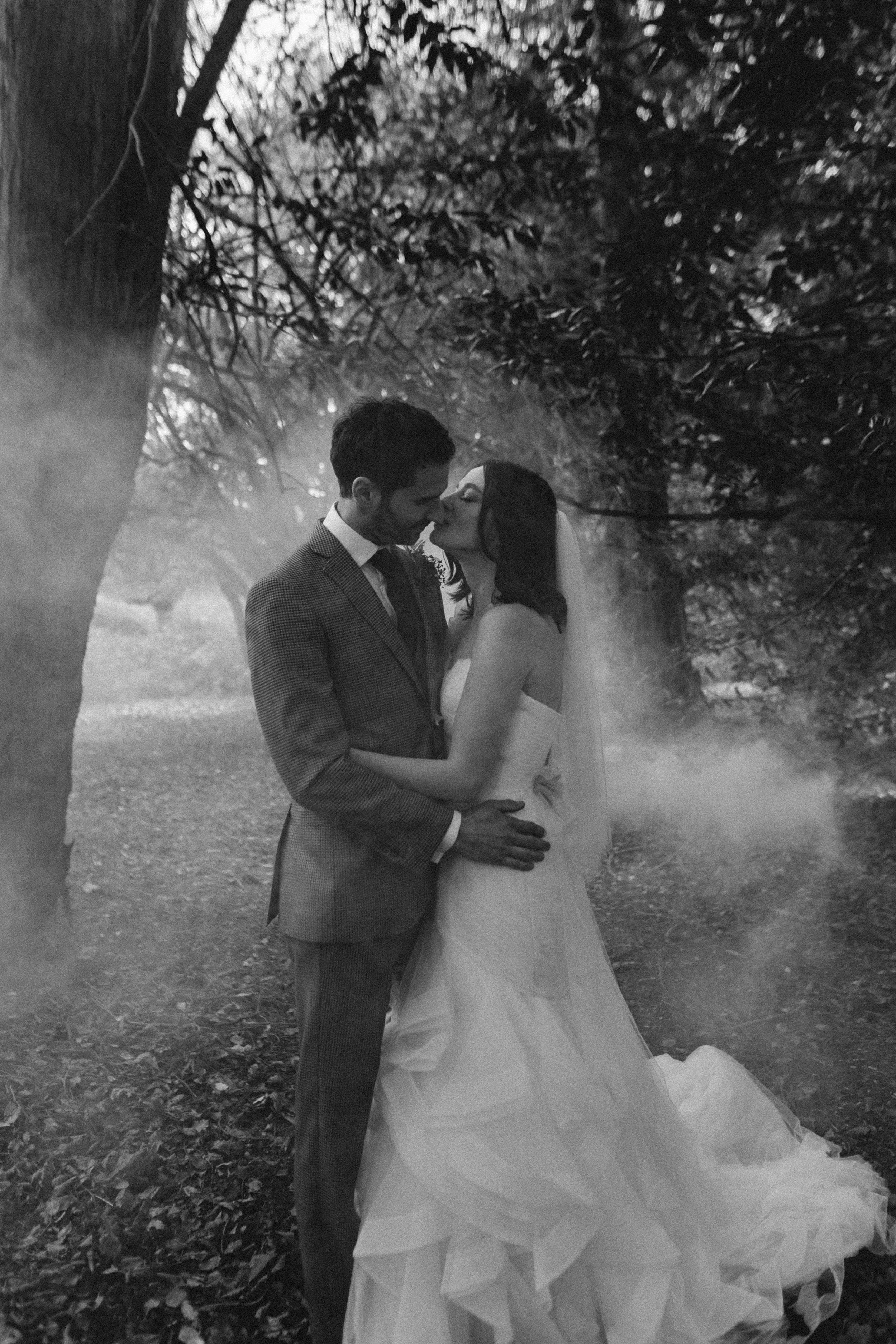 A couple in wedding attire sharing a kiss in a wooded outdoor setting, black and white photo.