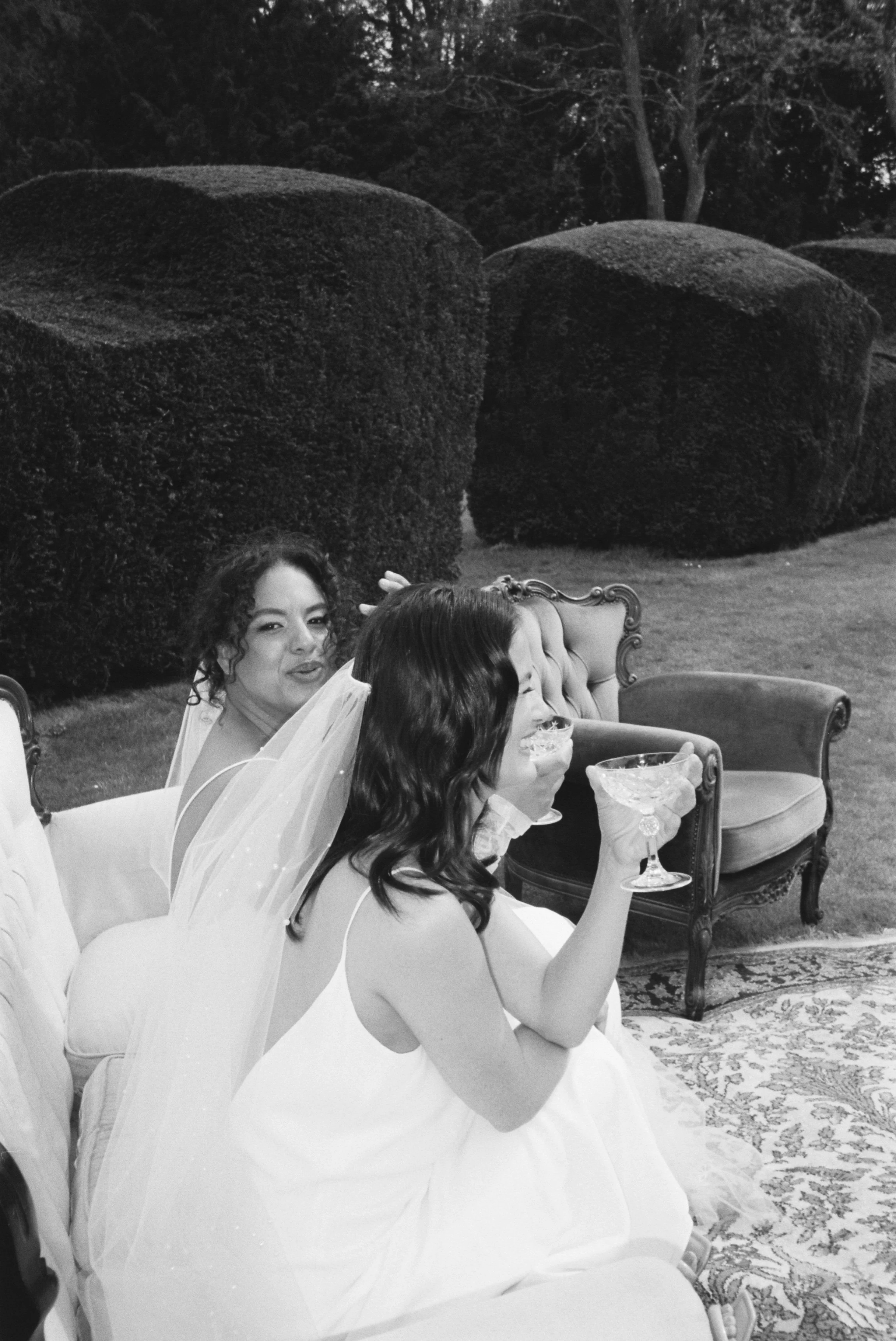 Two women in wedding dresses sit on vintage furniture outside, holding cocktail glasses, with manicured bushes and trees in the background.