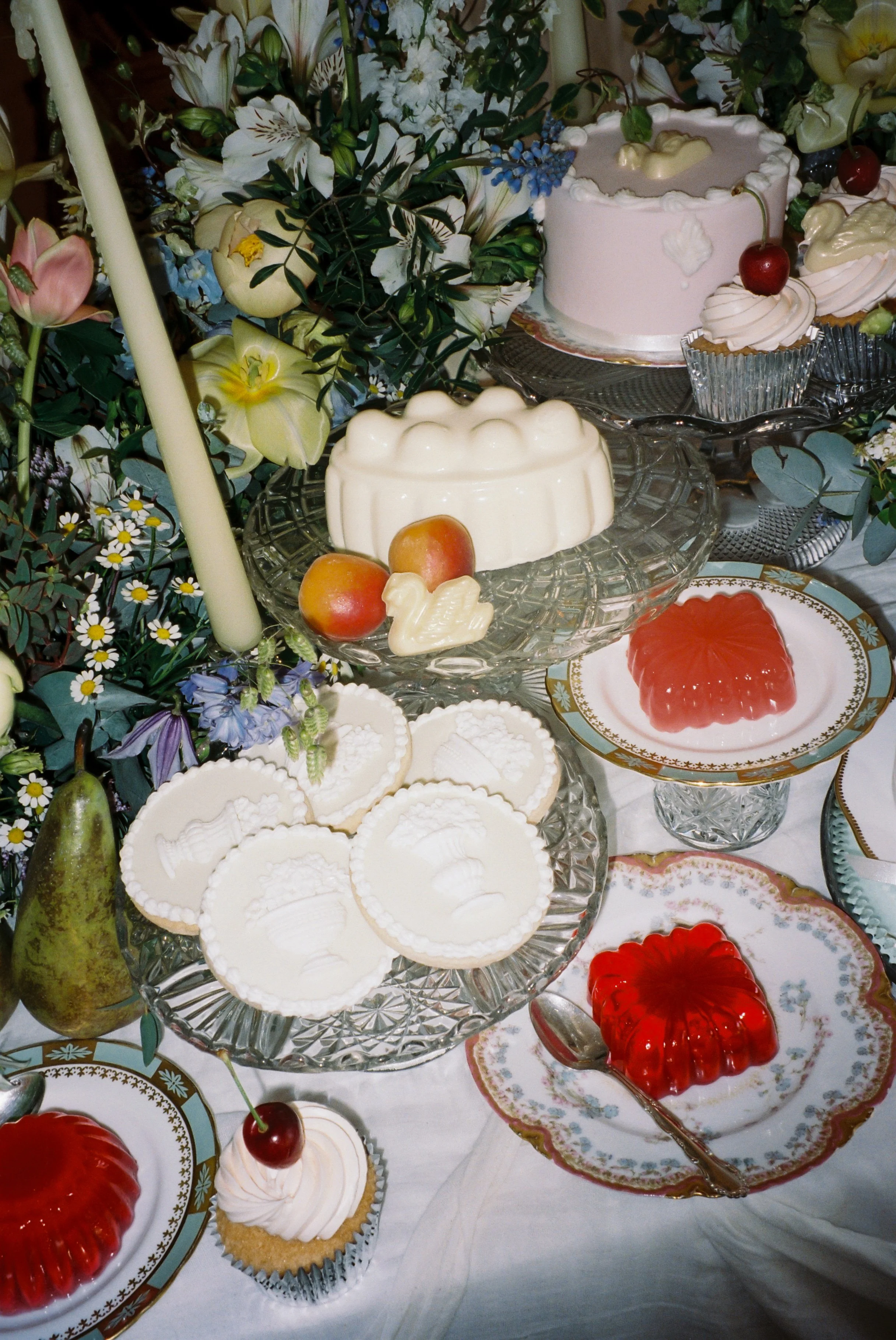 An elegant table setting with assorted desserts including jellies, cupcakes with cherries, a cake, and decorative plates, surrounded by flowers and greenery.