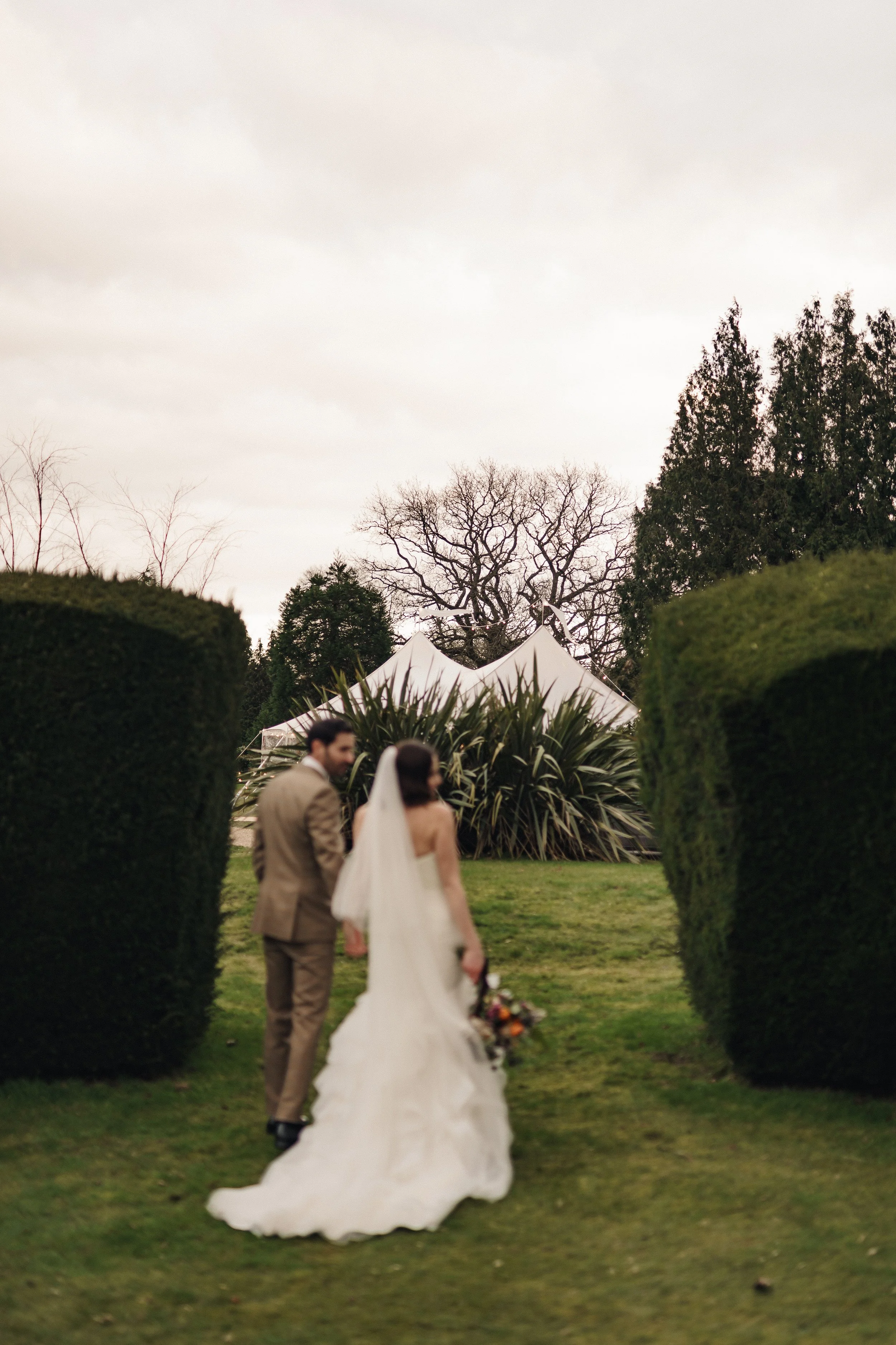 A bride and groom walking hand in hand outdoors, with a white tent, green bushes, and leafless trees in the background.
