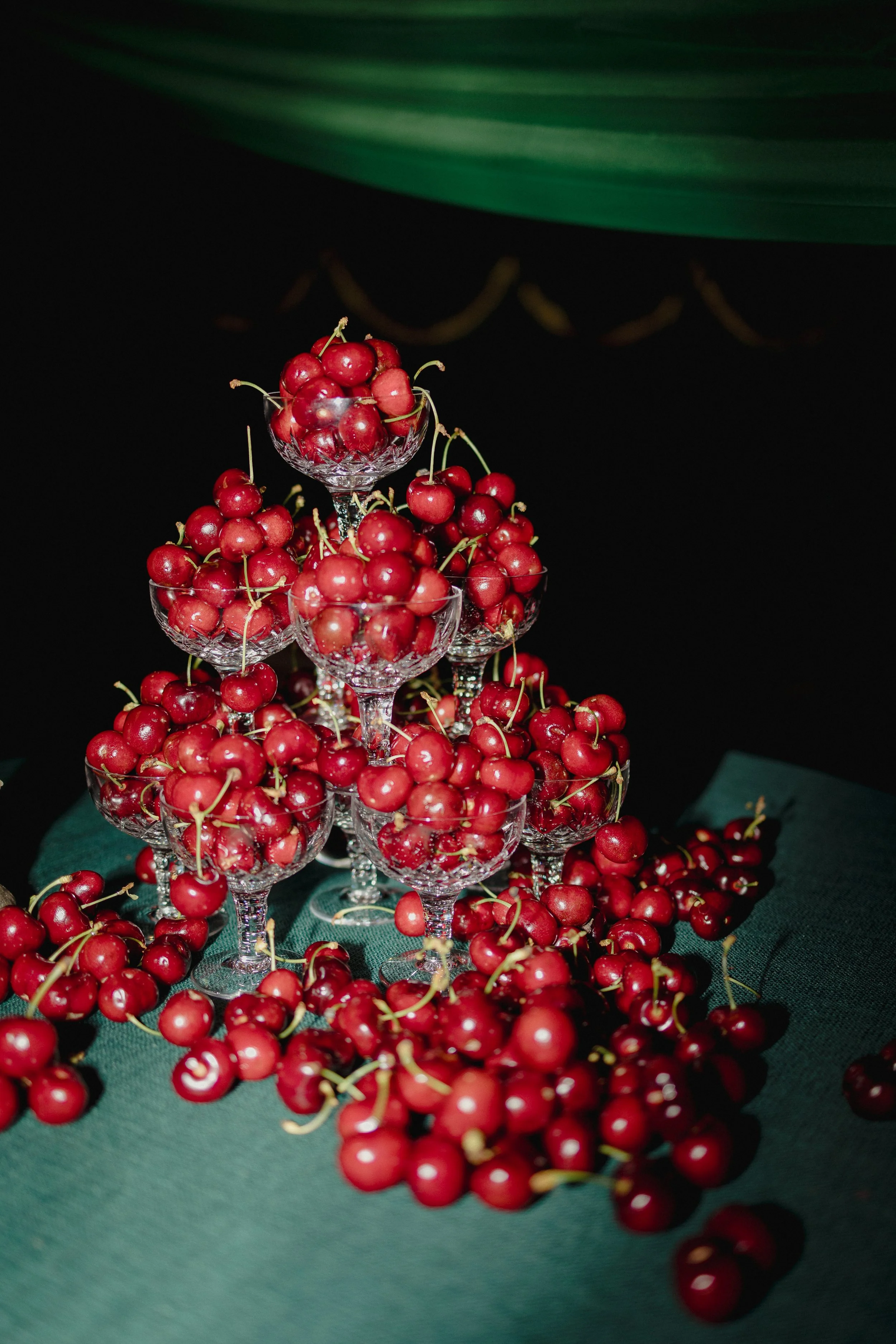 Glass dessert cups filled with red cherries arranged in a pyramid, with cherries scattered on a green cloth beneath.