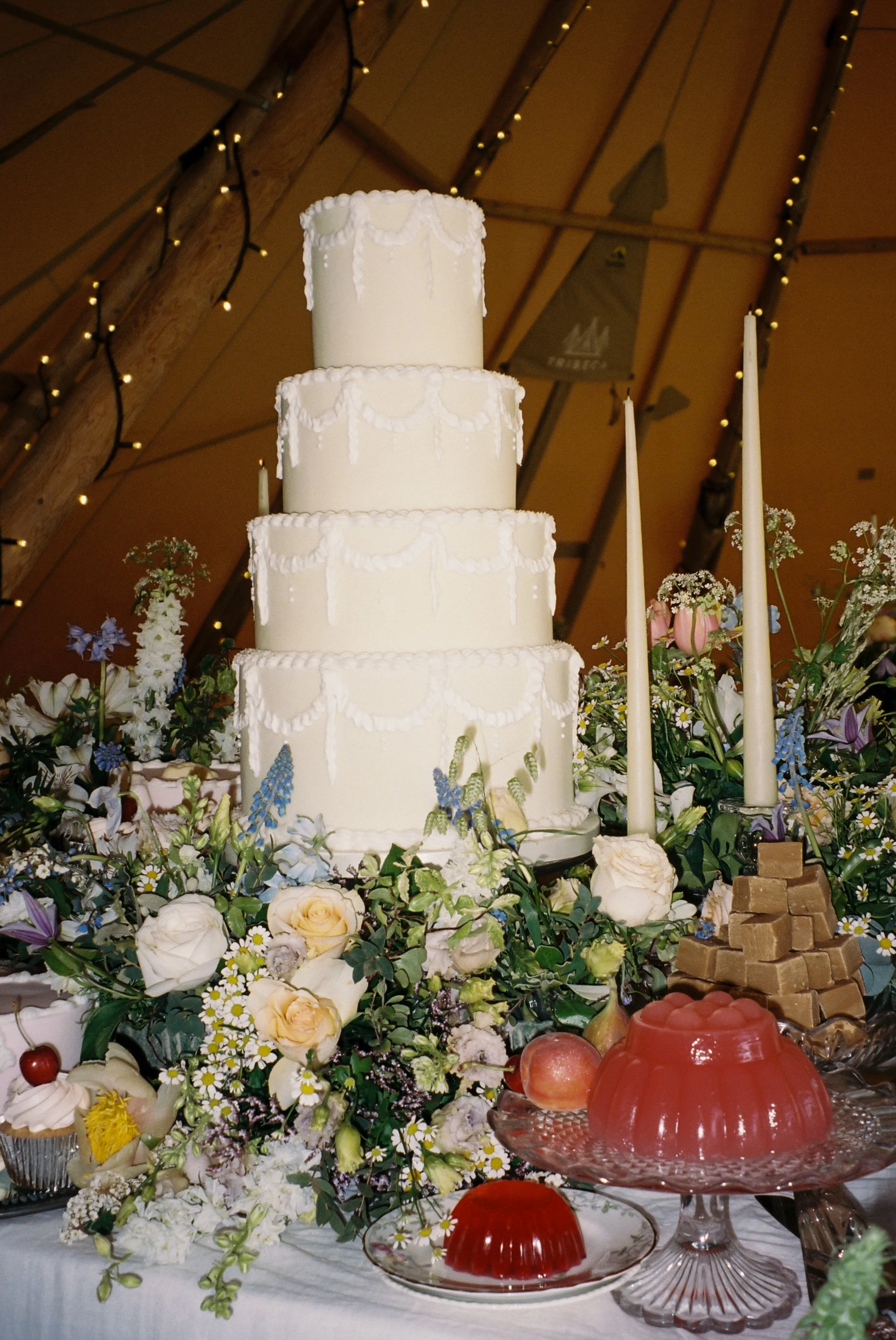 A tall, four-tier white wedding cake decorated with white icing swags, surrounded by flowers, candles, candies, and fruits on a decorated table.