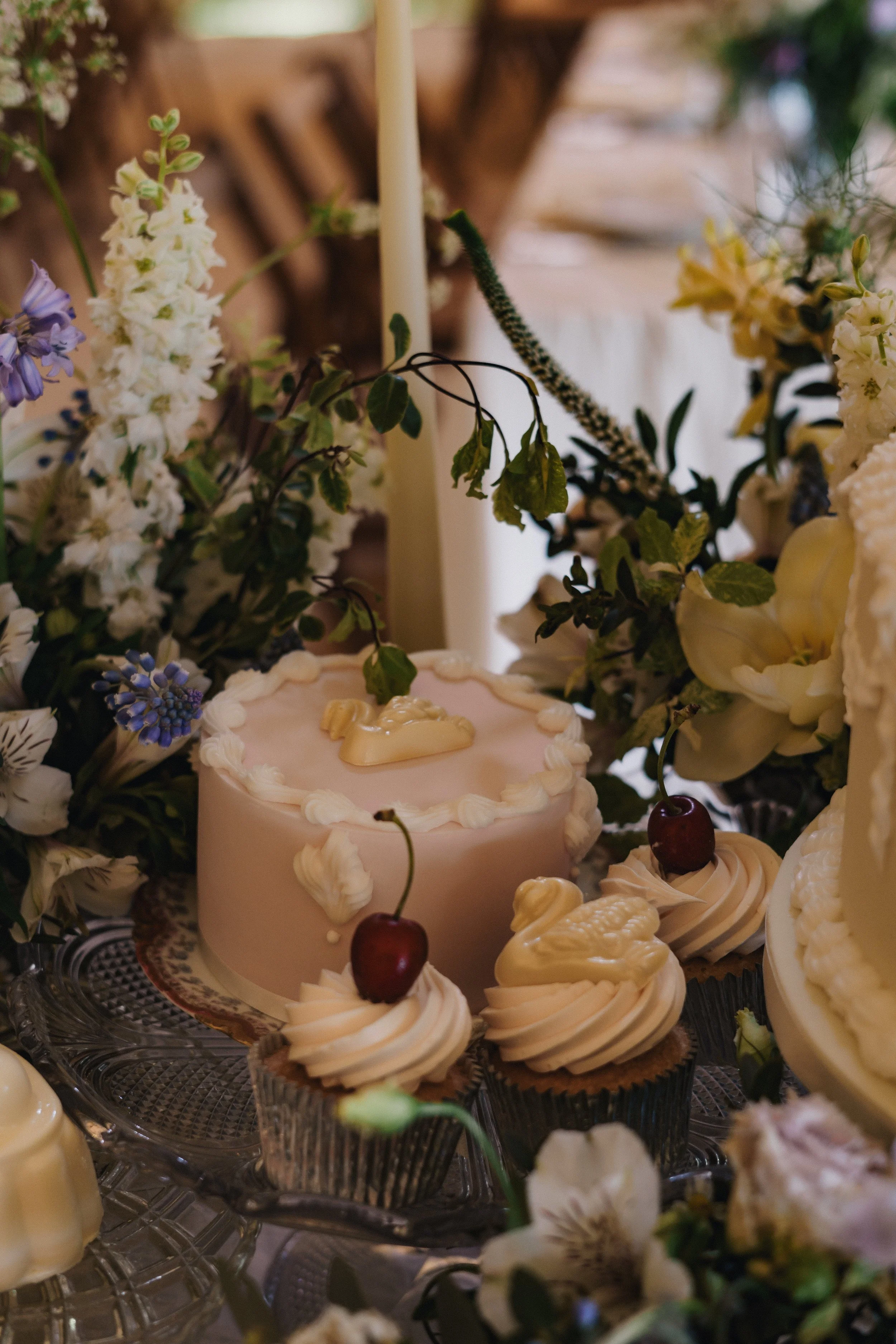 A pink cake with a white figurine on top, surrounded by cupcakes topped with cherries, on a glass tiered cake stand, decorated with flowers and greenery.