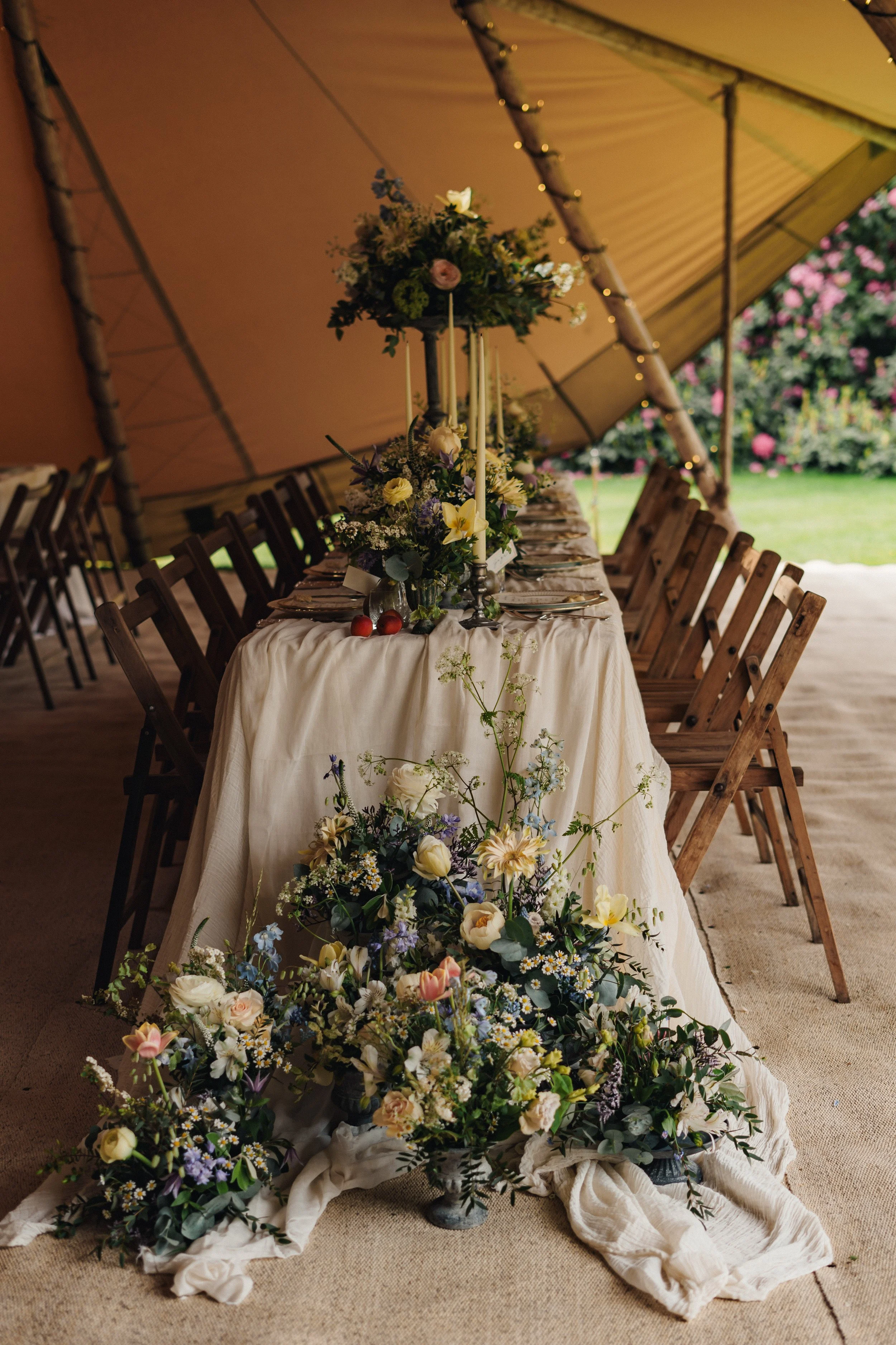 Decorative table setting for a wedding or celebration inside a large tent, with floral arrangements, wooden chairs, and outdoor greenery visible in the background.