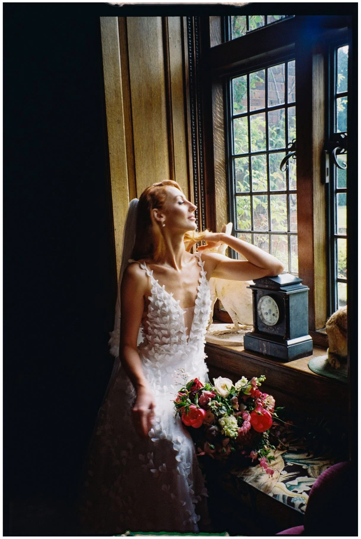 A woman in a wedding dress standing by a window, holding a bouquet of flowers, with sunlight illuminating her face.