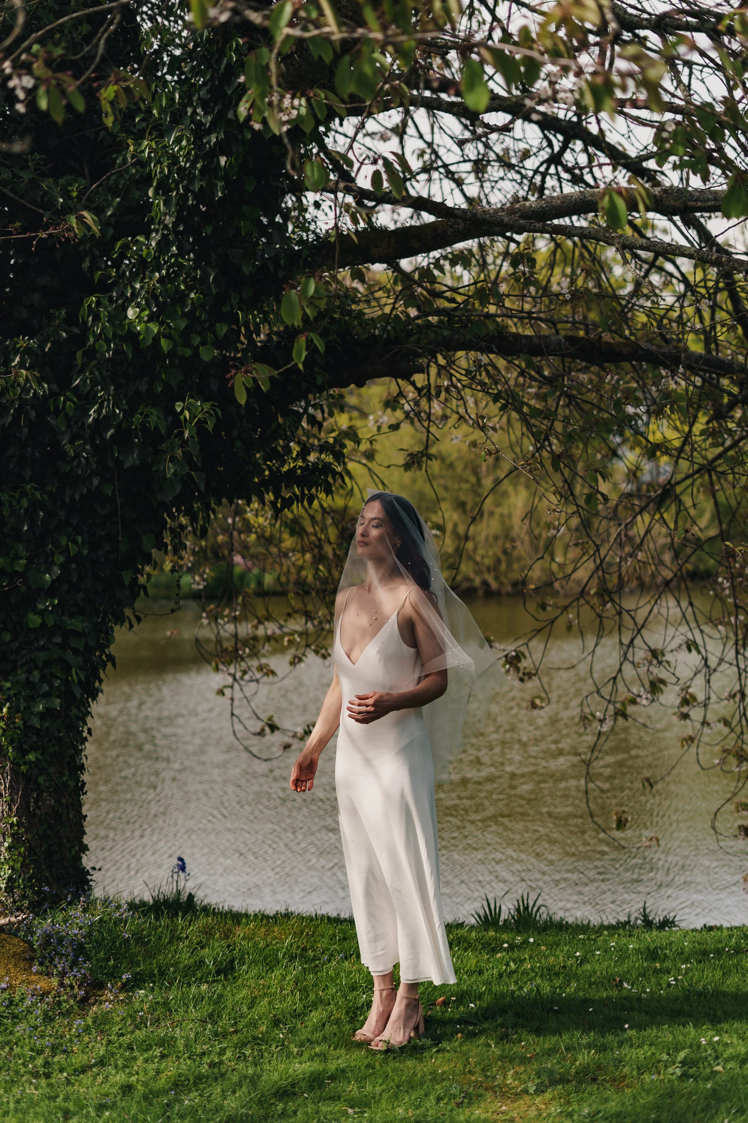 A woman in a white dress and veil standing near a lake with trees and greenery in the background.
