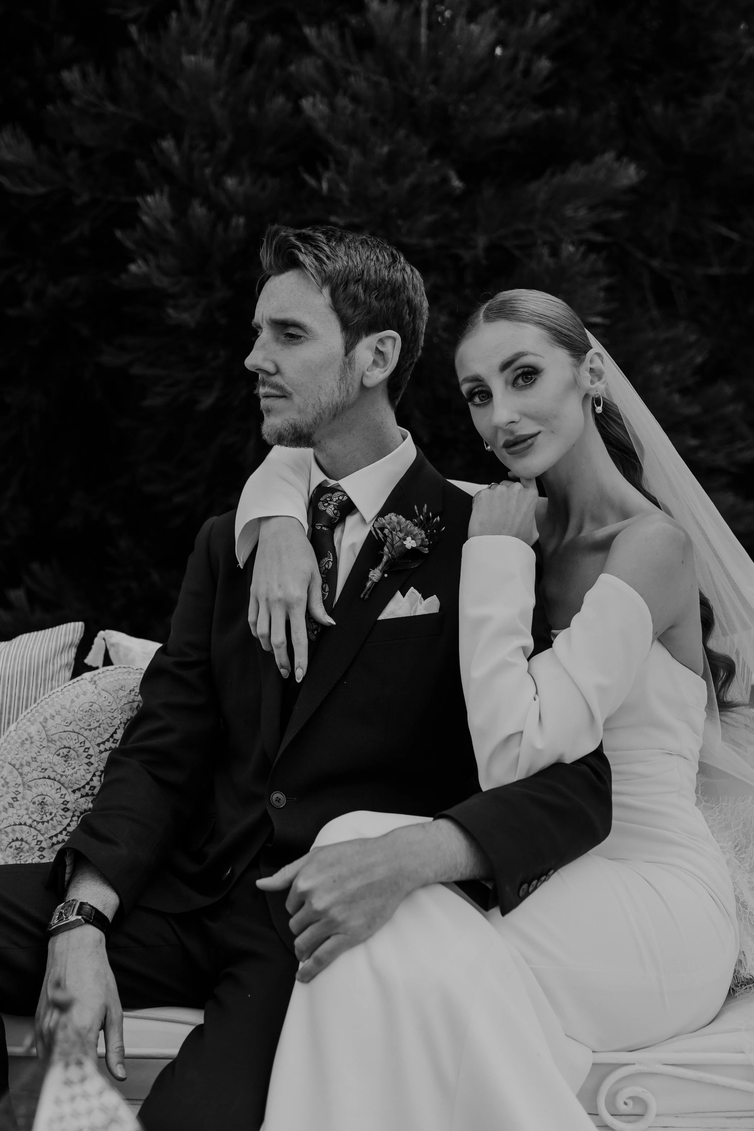 Black and white photo of a bride and groom sitting together outdoors. The groom wears a dark suit with a white shirt and tie, while the bride wears a strapless wedding dress and veil. They are sitting on a bench with pillows, with trees in the backgr