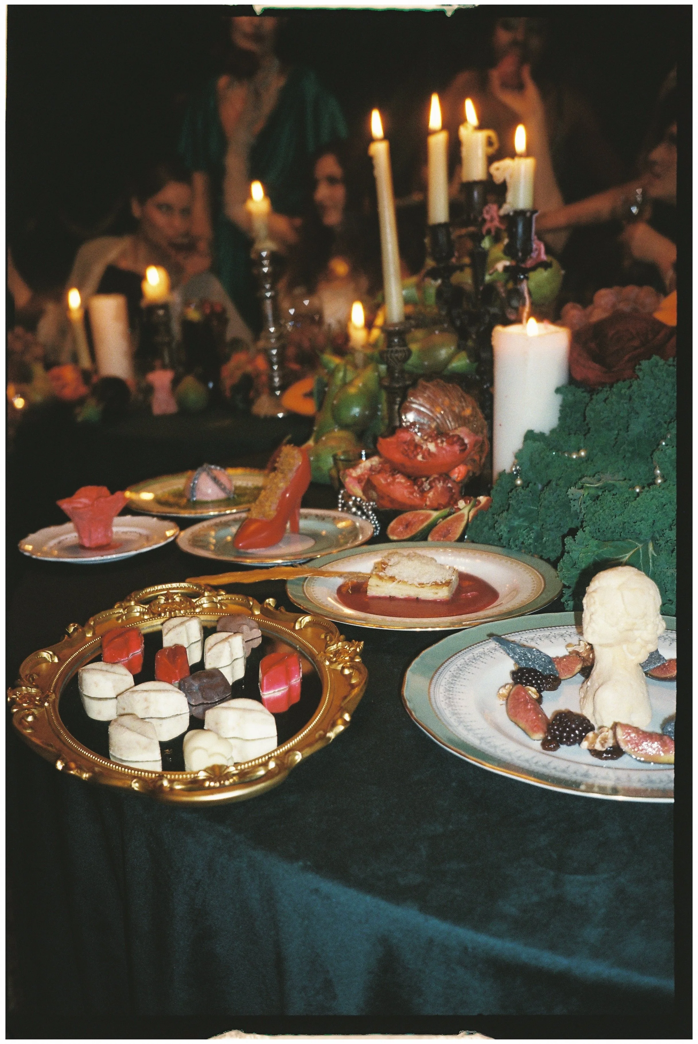 Decorative table setting with various desserts, including candies, a slice of pie, and a sculpted dessert, set against a dark background with candles and people in the background.
