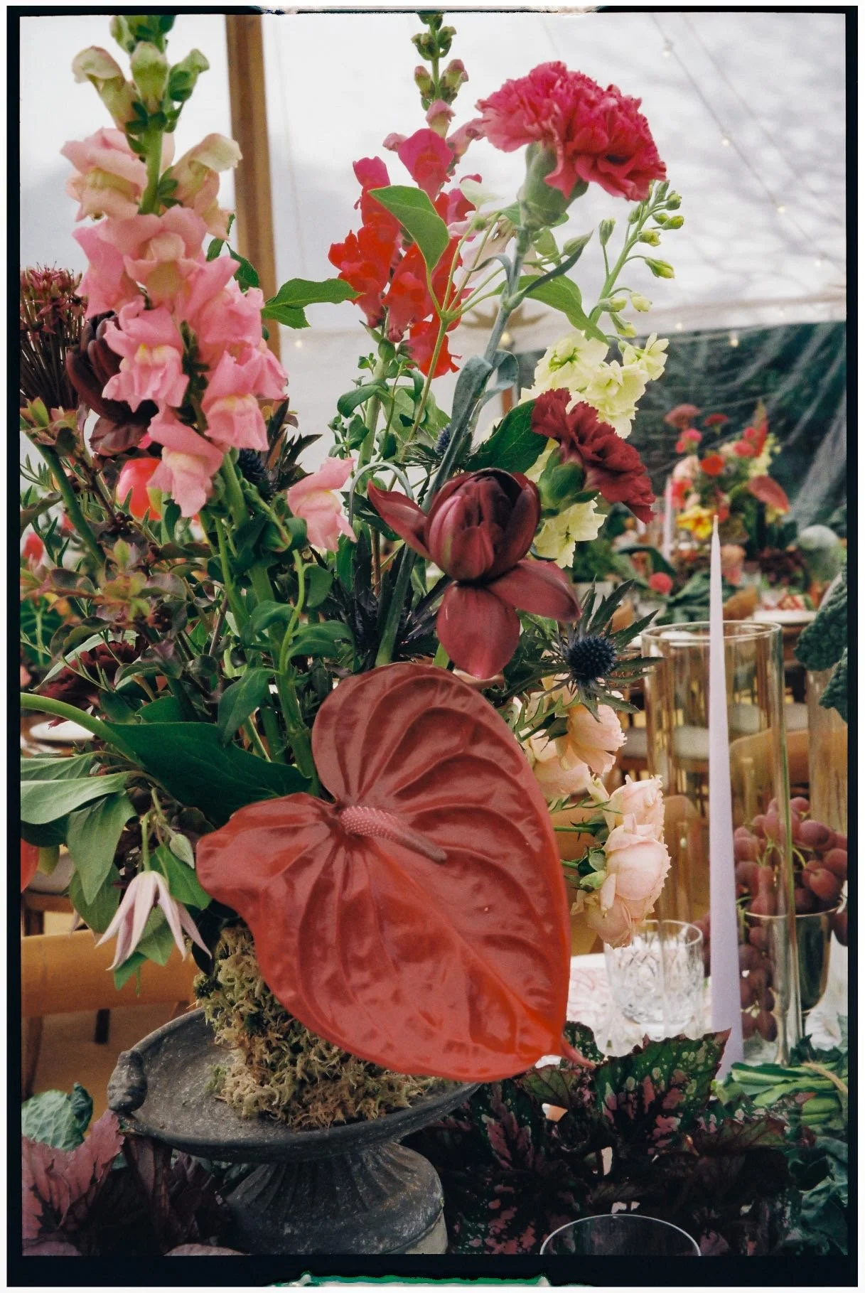 A vibrant floral arrangement featuring pink, red, and beige flowers with green leaves, placed in a gray stone pedestal vase, with tall yellow candles in glass holders in the background, on a decorated table.