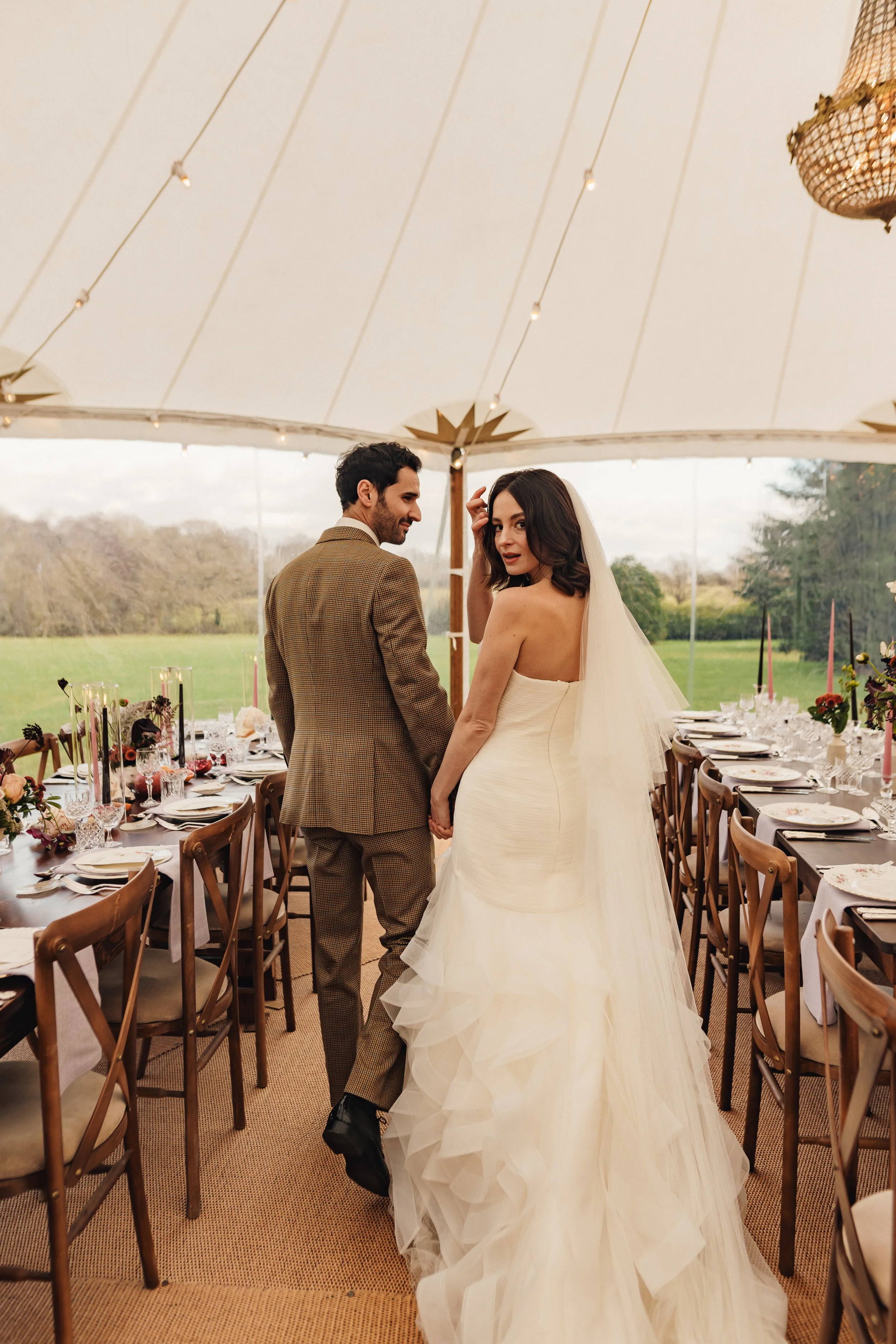 A bride and groom holding hands inside a decorated wedding tent with long dining tables set for a celebration.