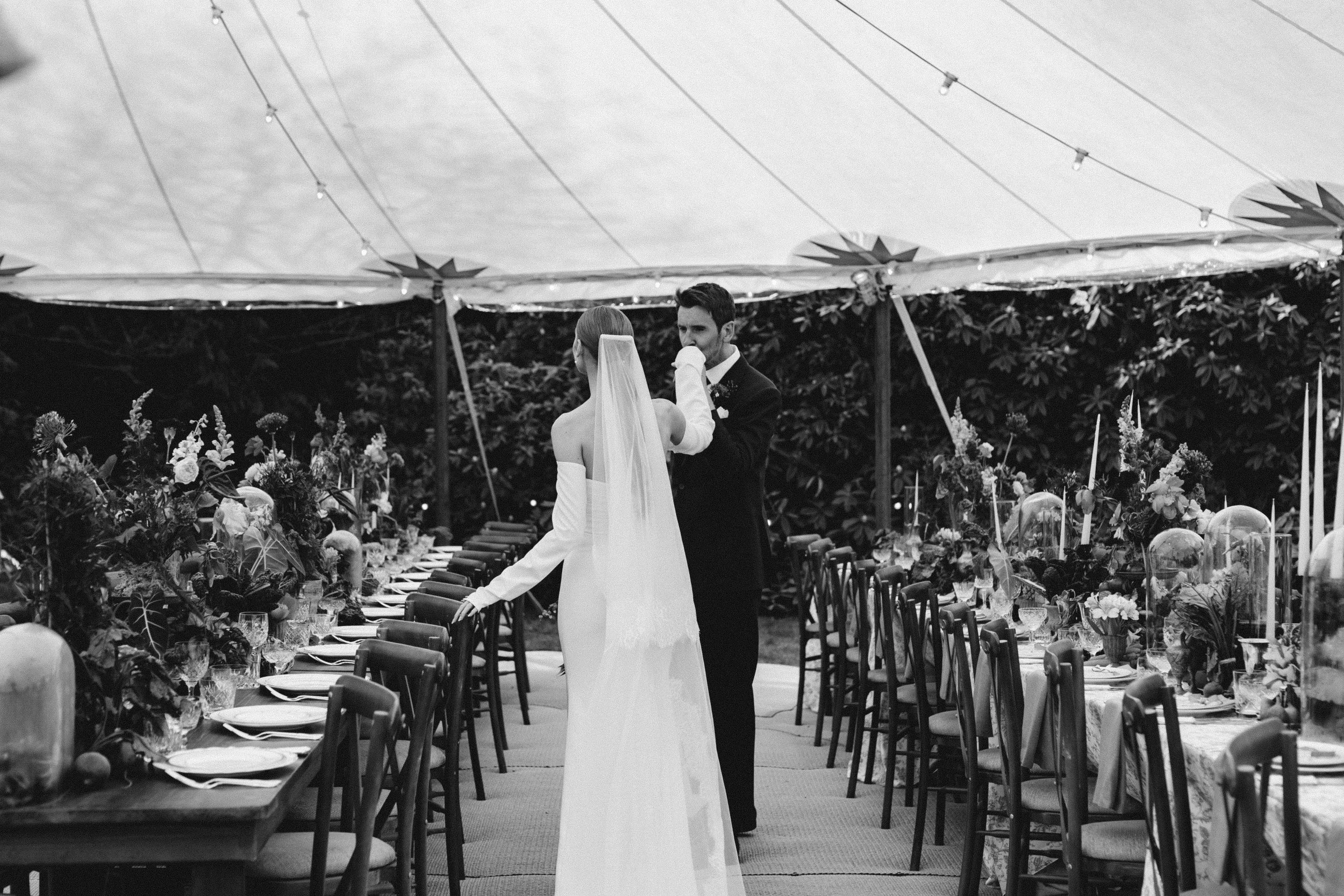 Black and white photo of a bride and groom in wedding attire under a canopy, standing near a long decorated dining table with chairs and floral decorations.