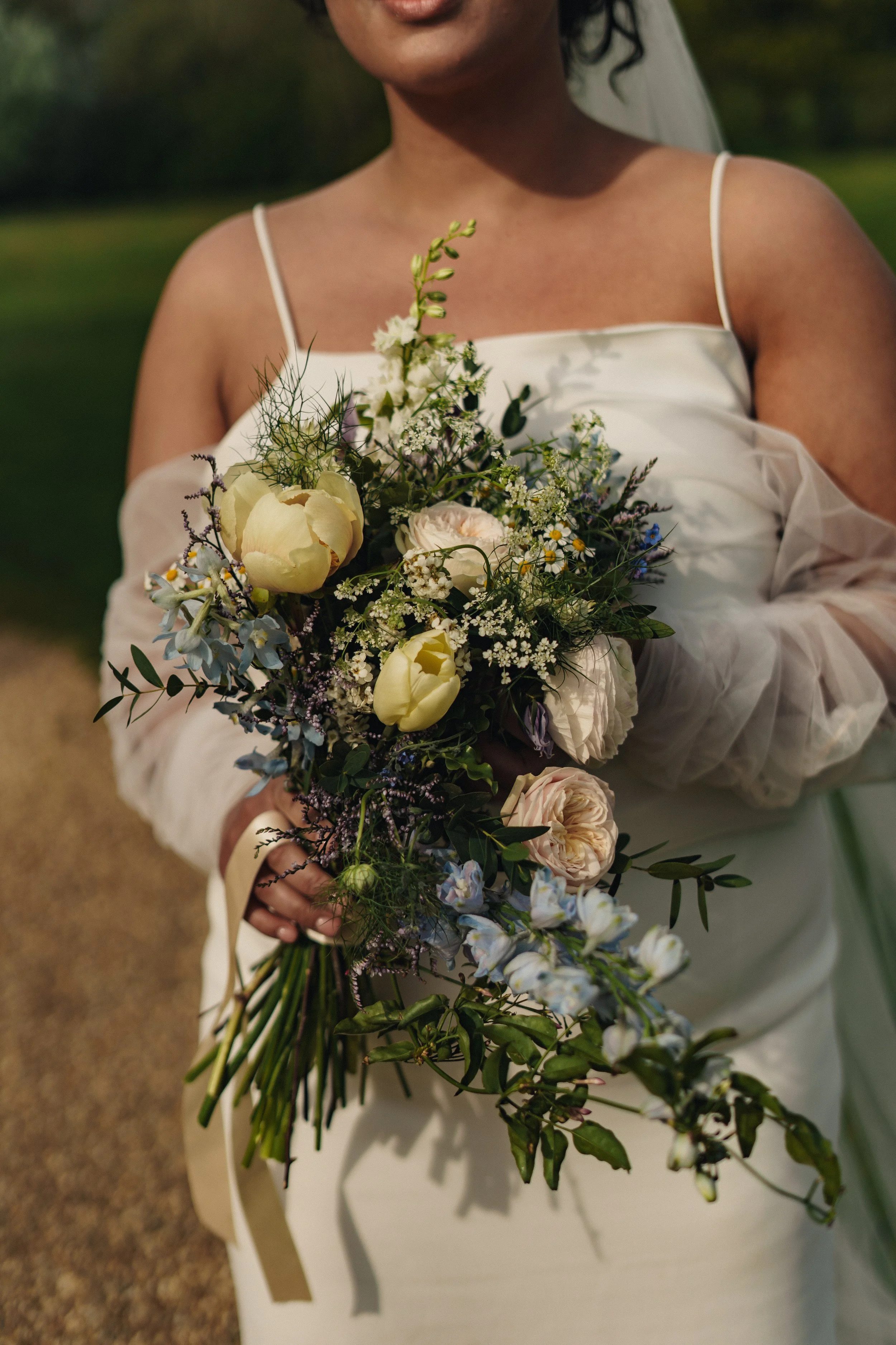 A person in a white dress holding a bouquet of mixed flowers, including peonies, roses, and greenery, outdoors in natural light.