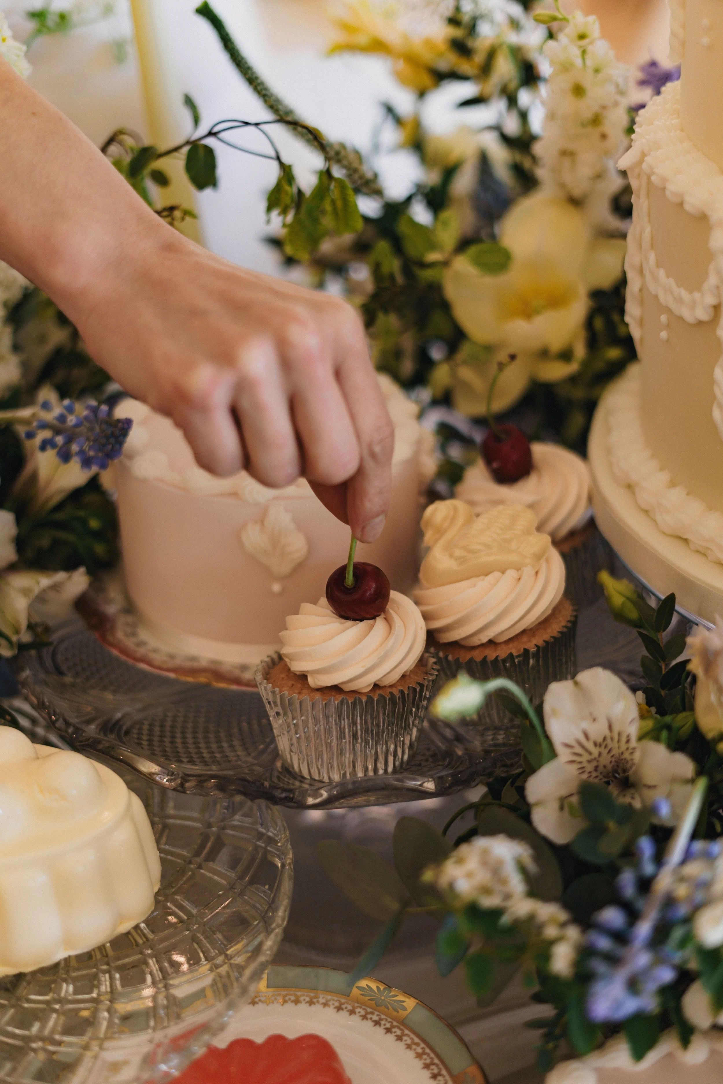 A person placing a cherry on top of a cupcake at a cake display with flowers and other desserts.