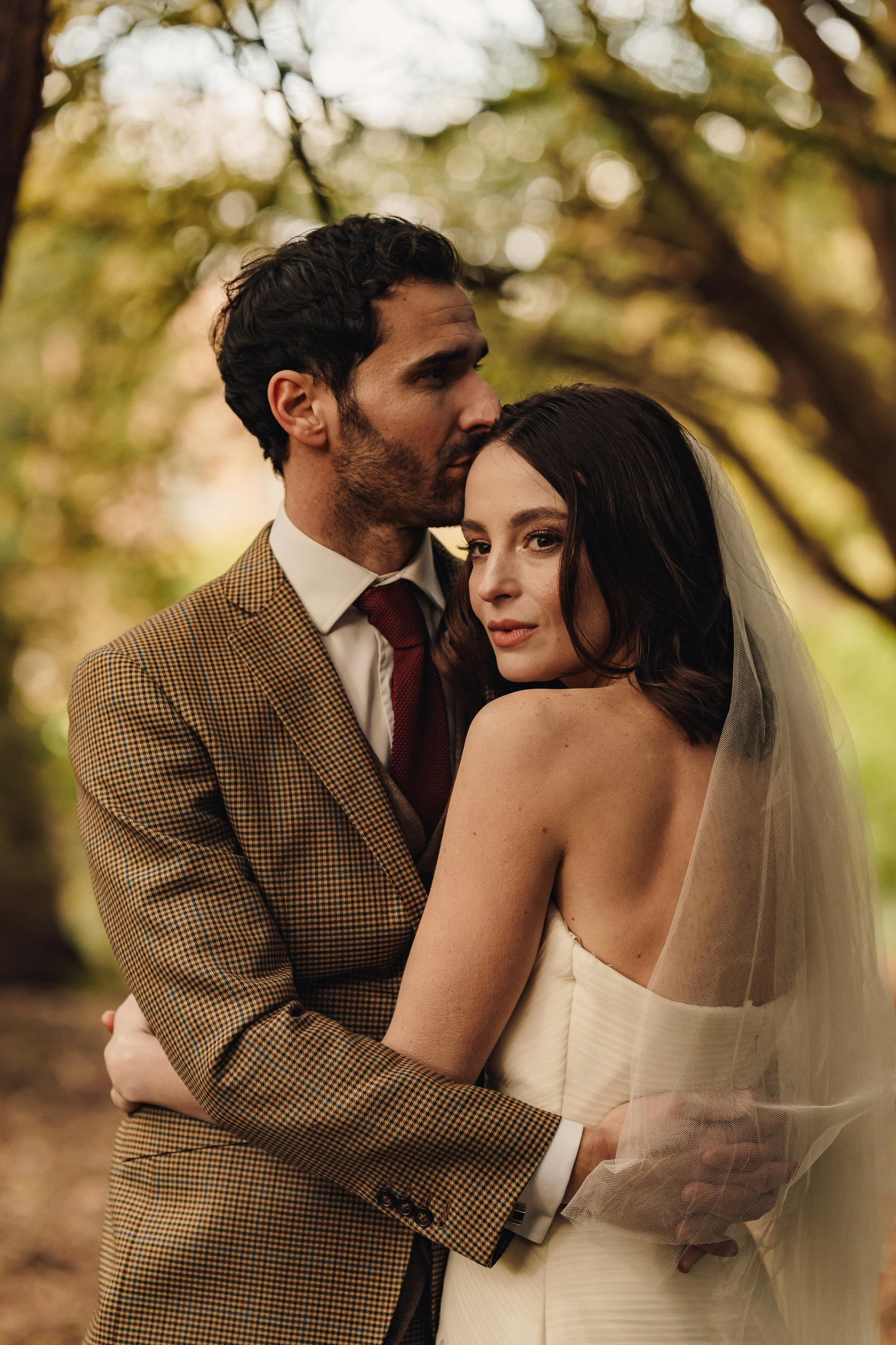 A man and woman embrace outdoors during autumn, the man in a checkered brown jacket and the woman in a strapless white dress with a veil.